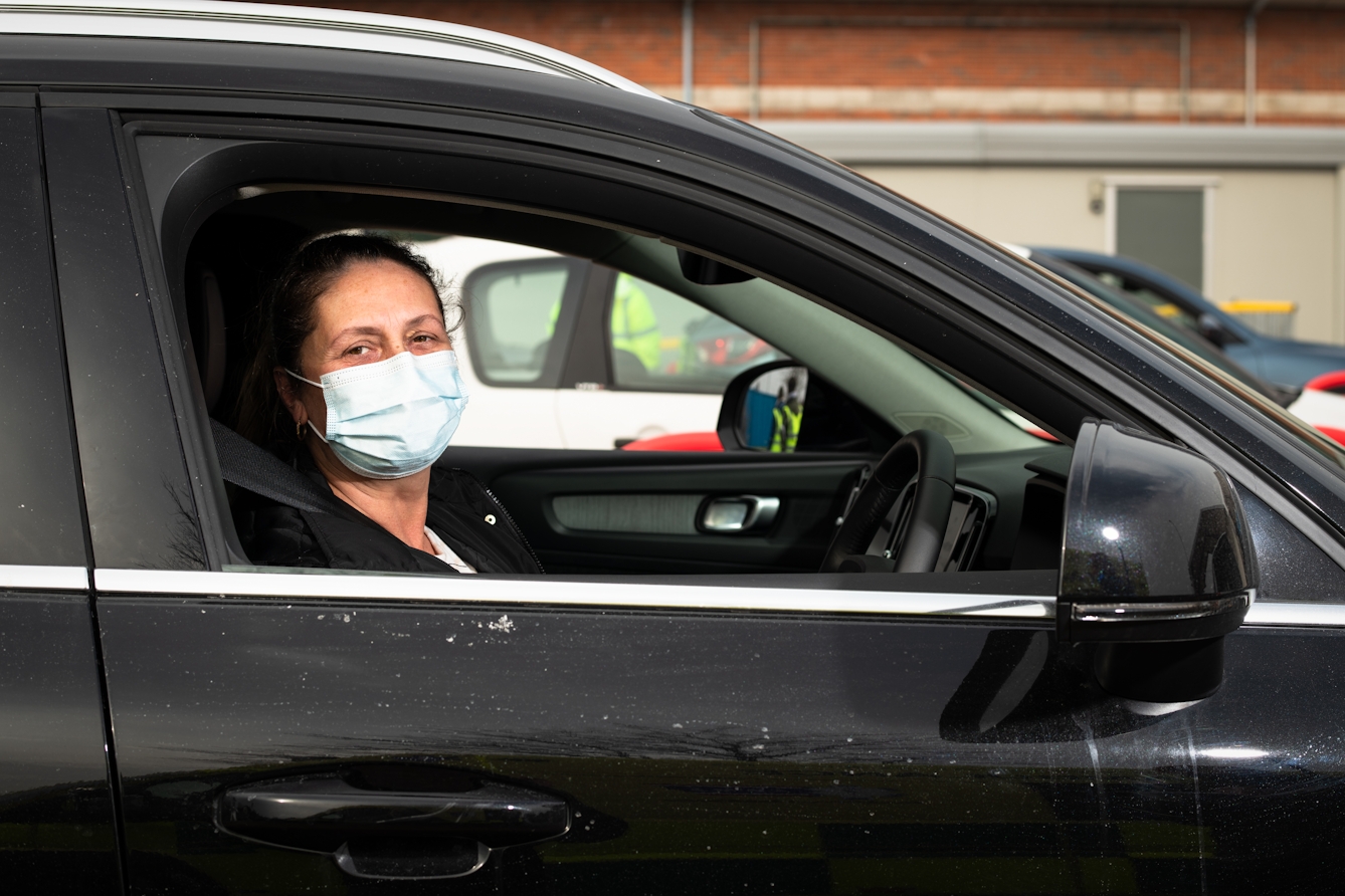 Photographic flash lit portrait through the open side window of a stationary car. The car is black and the front of the car is facing to the right. Sat in the driver's seat is a woman, looking to the camera. She is wearing a blue medical face covering. In the background is the out of focus scene of a building and other cars.