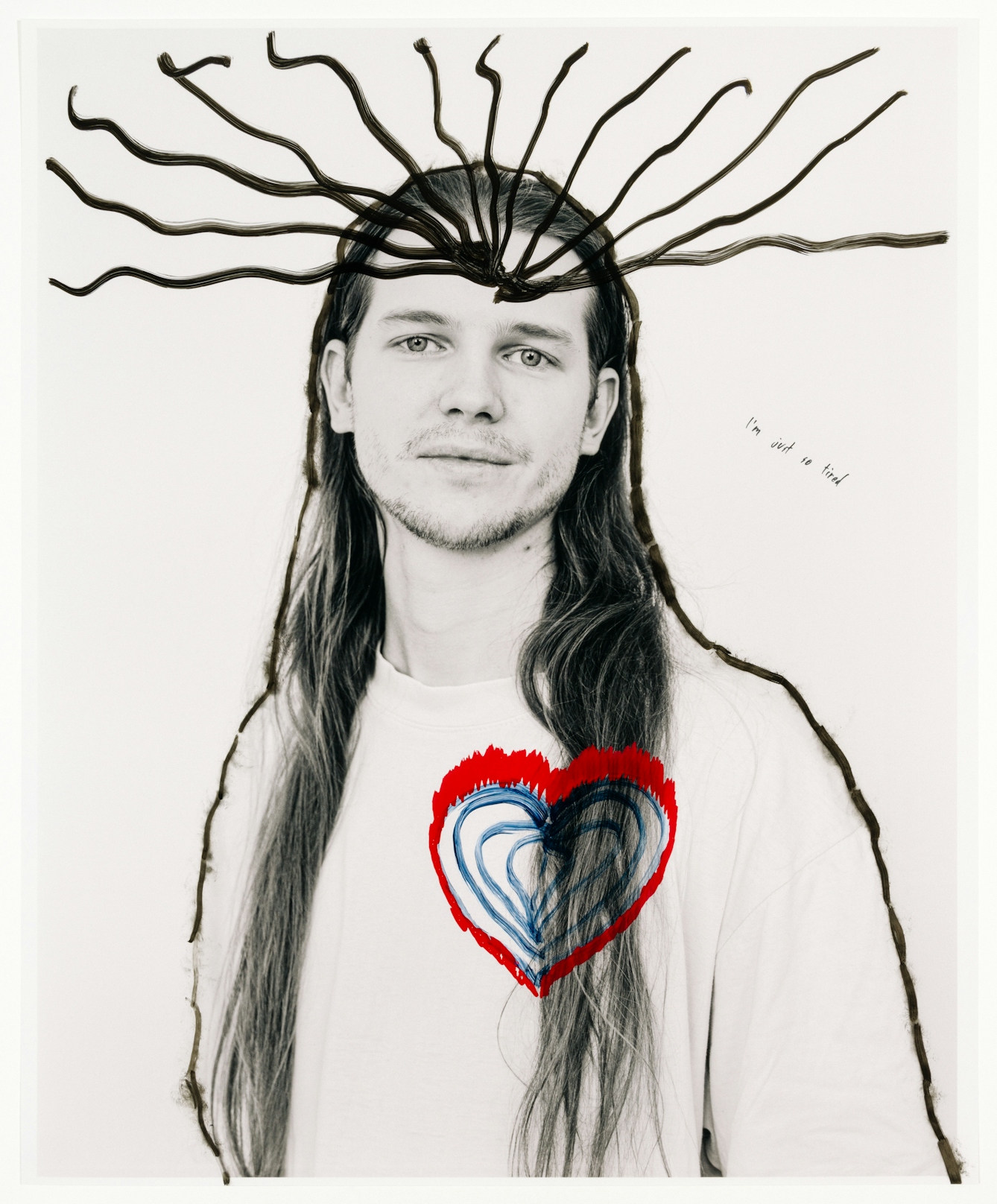 Head and shoulders photograph of a man with long hair staring at the viewer.  His body has been outlined with a black marker pen; hand drawn on the photograph.  A series of lines emanate from the man’s head.  A blue and red heart shape has been drawn on his chest.  In small handwriting, the words “I’m just so tired” have been written to the left of his portrait.