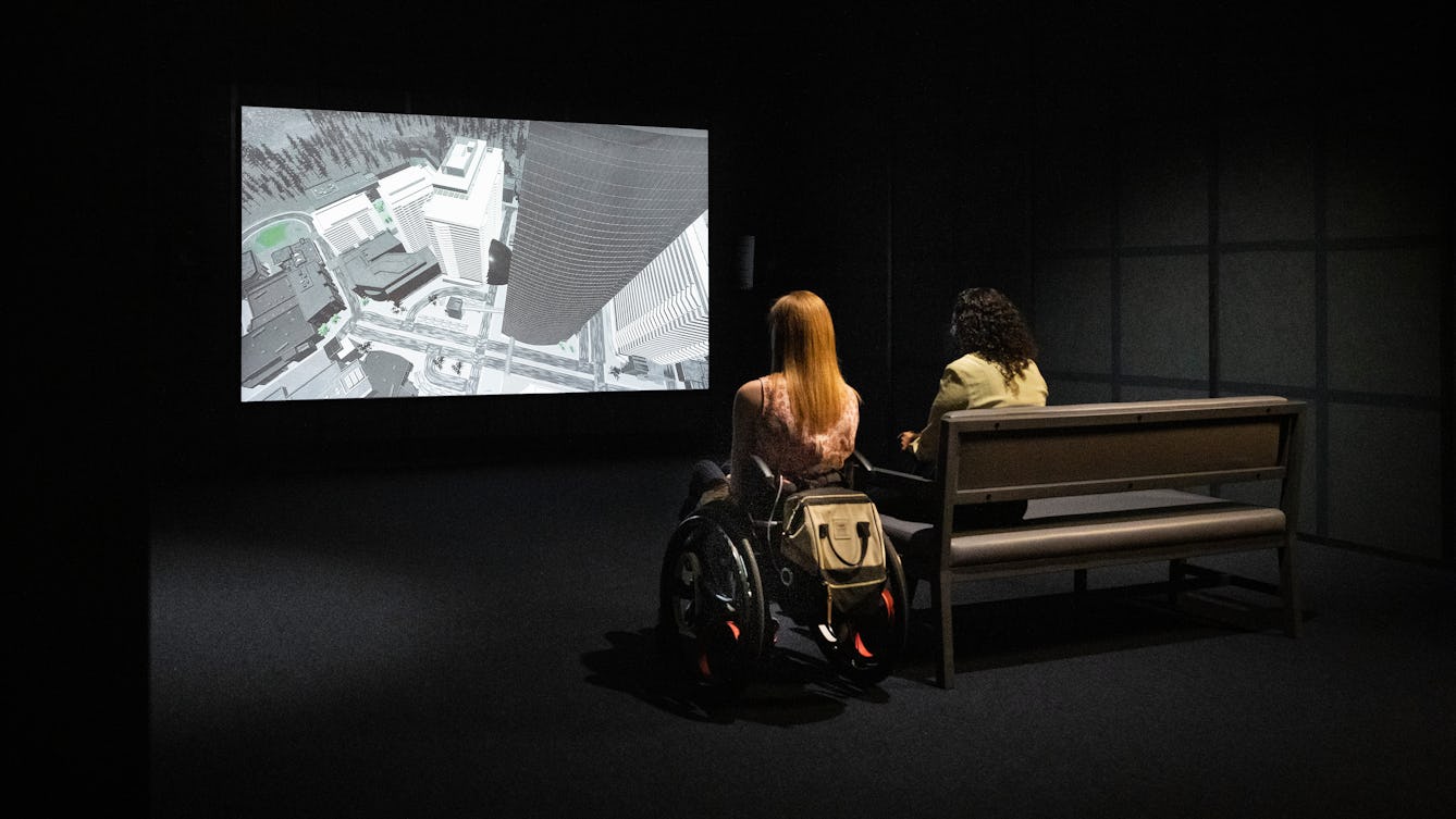 Photograph of a dark exhibition space featuring a projected video on a large screen. Three visitors are watching the film. 