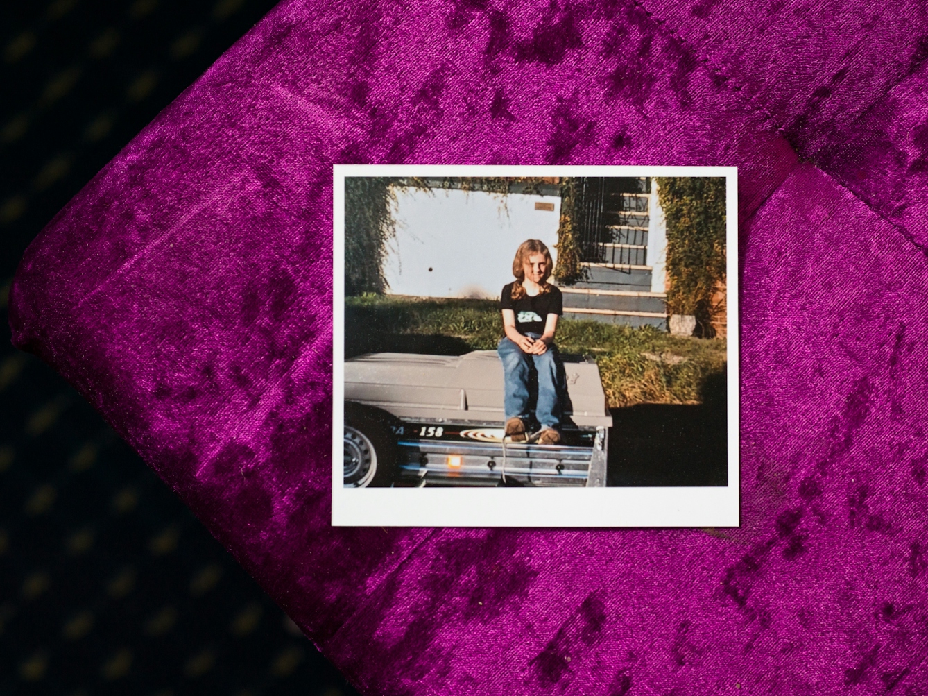 Photograph of print of a family photo of a young girl outside, sitting on the top of a car trailer. She is wearing jeans and a black t-shirt and is smiling to camera. Behind her is a grassy bank next to the road and the front white garden wall, gate and steps of a house. The print is lying on top of a purple velvet fabric which is part in shade, part in sunlight.