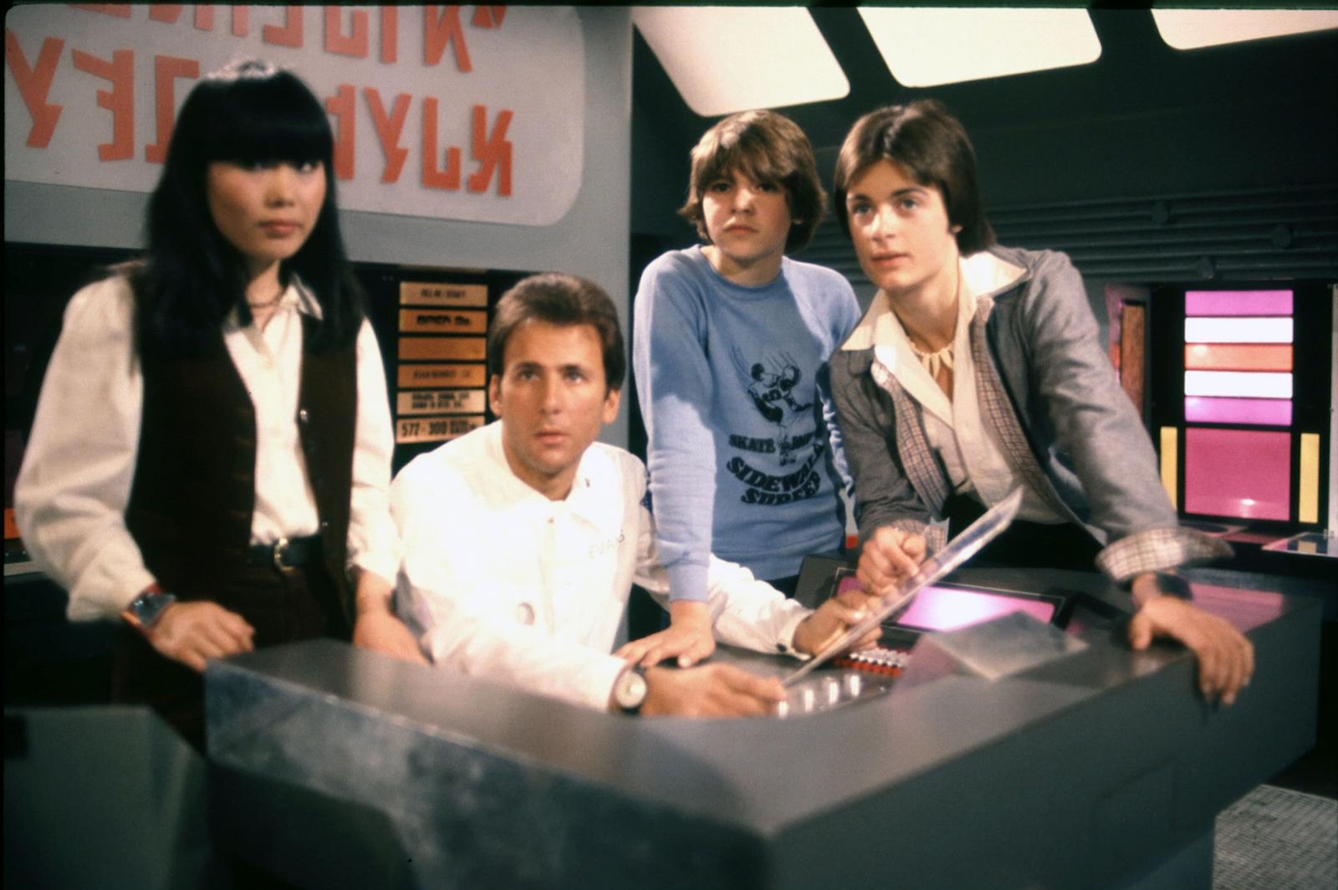 Four young people clustered around a futuristic control desk behind which there is alien lettering.