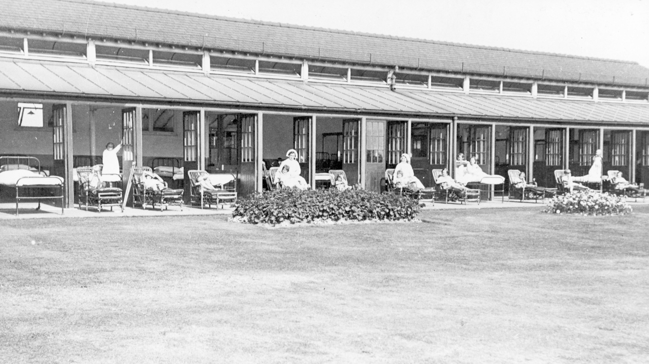 Black and white photograph of a long single storey building used as a hospital ward. Multiple doors are open showing a row of beds inside and children attended by murses sitting on sun loungers in front of the building, looking out across a well manicured lawn.