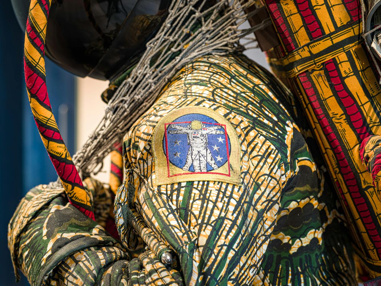 Photograph of a close-up detail of the shoulder of a life-size artwork of a figure resembling an astronaut, carrying a large net containing assorted objects. On the shoulder is an embroidered badge showing an astronaut with arms and legs outstretched within a blue circle.