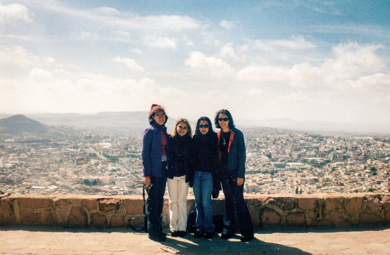 Photograph of a group of four young women standing by a low wall, behind which a vista of Mexico City extends into the distance.