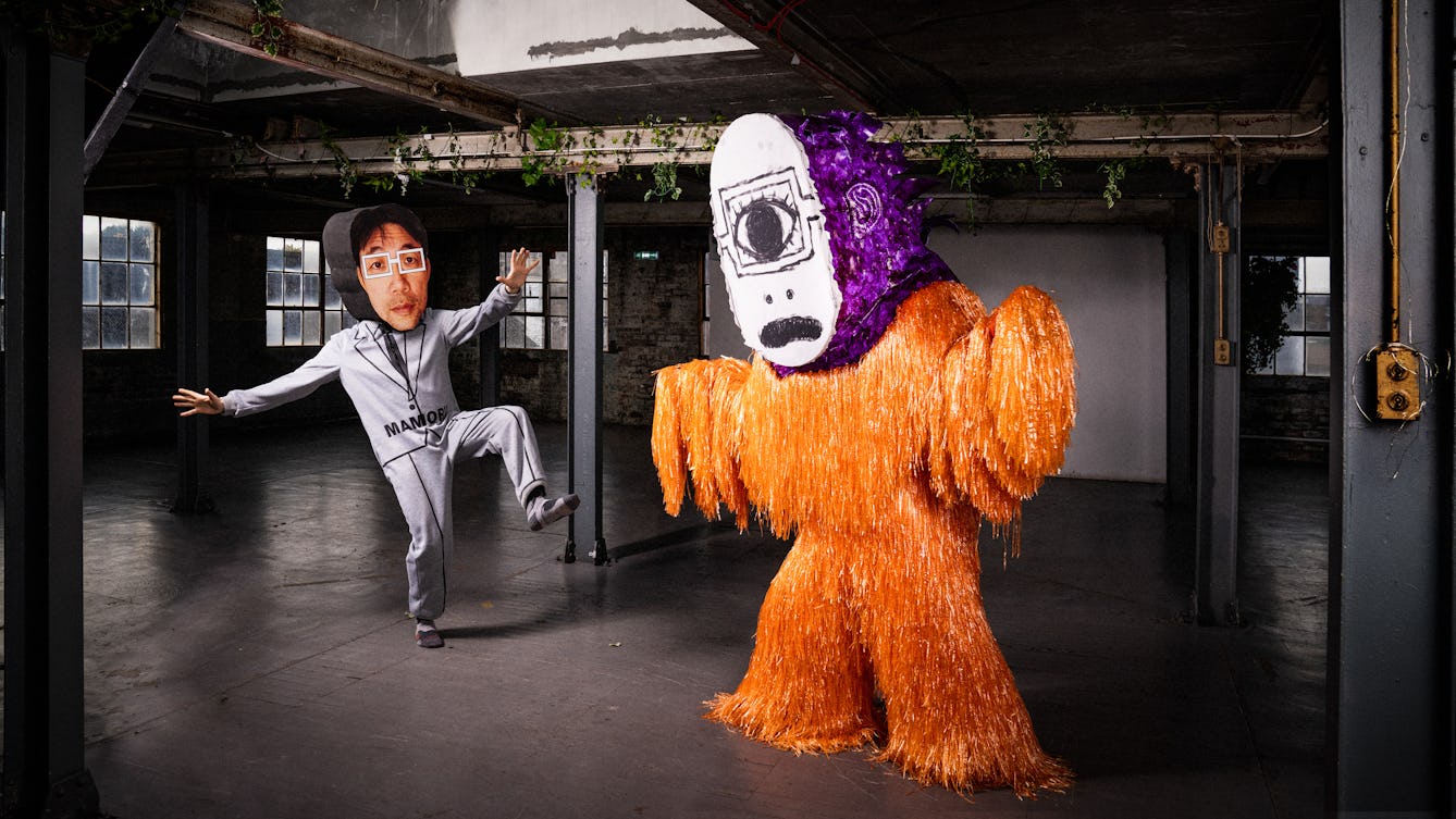 Two people performing in costume in a warehouse. A furry orange cyclopes with one large eye and a purple head is looking down at Mamoru Iriguchi who is photographed mid-movement, with one leg in the air and hands out. On his head is an oversized 2D double-sided face with square glasses, he is wearing a grey costume with a drawn on suit and tie. "Mamoru" is printed in large black letters on the suit jacket. 