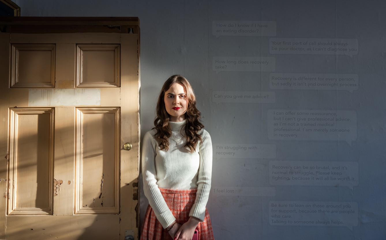 A photograph of Emily Basforth standing in front of a grey wall. She is wearing a cream jumper, an orange plaid skirt and red lipstick. She has long brown wavy hair. To the left, there is a wooden door. To the right, there is a text message conversation superimposed on the wall. 

The text messages read: 
'How do I know if I have an eating disorder?'
'Your first port of call should be your doctor as I can't diagnose you'
'How long does recovery take?'
'Recovery is different for every person but sadly it isn't and overnight process.'
'Can you give me advice?'
'I can offer some reassurance, but I can't give professional advice as I'm not a trained medical profession. I am merely someone in recovery.'
'I'm struggling with recovery'
'Recovery can be so brutal, and it's normal to struggle. Please keep fighting, because it will all be worth it.' 
' I feel lost'
'Be sure to lean on those around you for support, because there are people who care. Talking to someone always helps.'