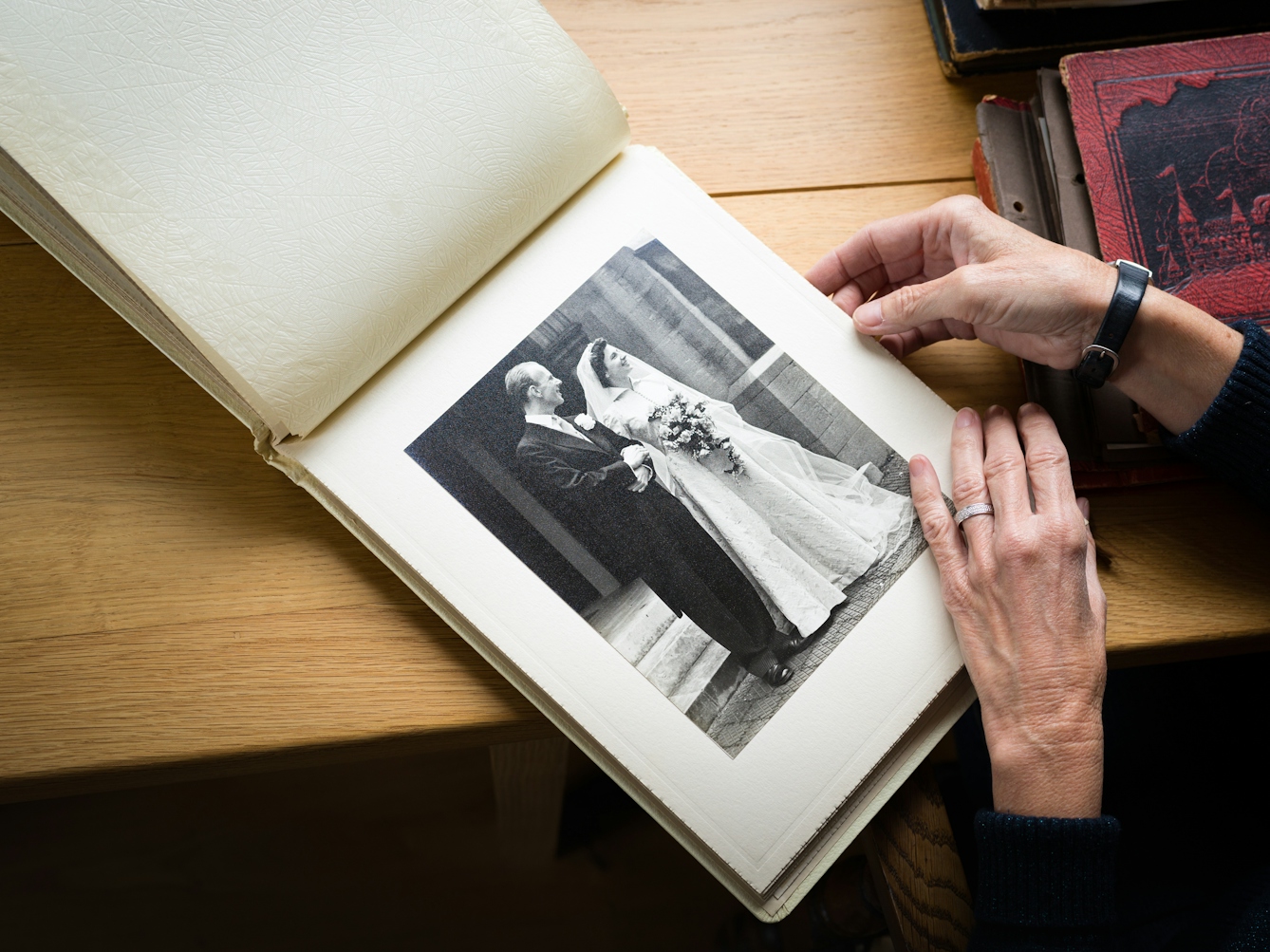 A photograph of an open photo album with a large photo of a man and a woman on their wedding day. A pair of hands lay across the bottom right of the wedding photo and on the left hand there is a silver ring. The album is on a wooden desk and to the right are piles of other photo albums.