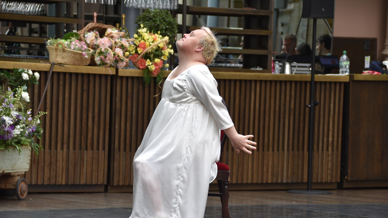 A performance showing Danny Smith in a white dress, posed on a chair. He is sitting side on, head raised and arms positioned behind him. 