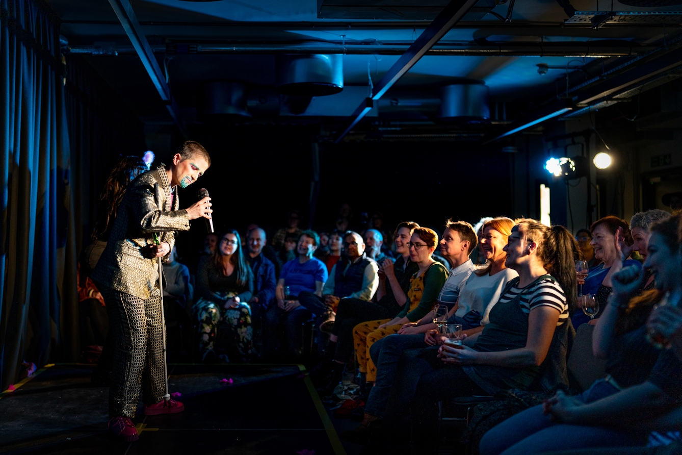 Photograph showing a performer on a stage in a small theatre. The performer is to the left of frame dressed in a shiny gold suit leaning forwards and speaking into a microphone in their right hand. In their left hand they are holding a white cane. To the right of frame is a seated audience.