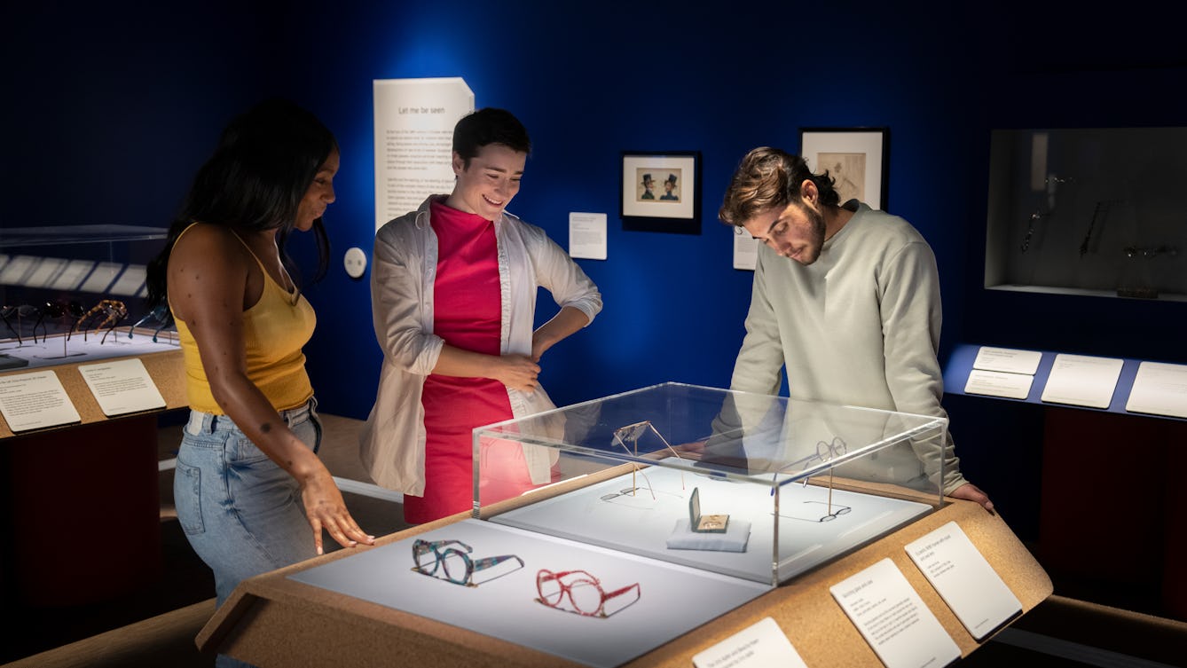 Photograph of three people exploring an exhibition. They are stood side by side at a display case which contains pairs of spectacles. Outside of the case are two pairs of spectacles which the visitors can touch and explore with their hands. The rest of the gallery behind them is dark blue.