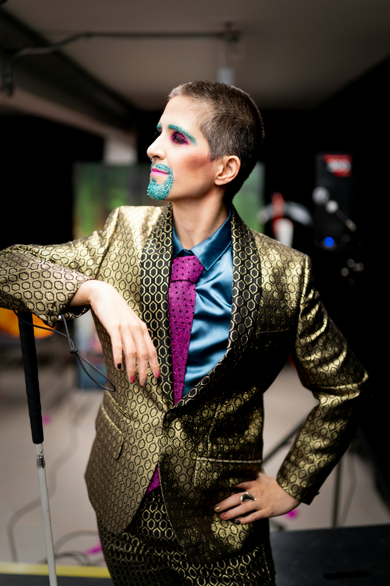 Photograph showing a performer backstage. The performer is dressed in a shiny gold suit. Their right elbow is resting on the handle end of a white cane. Their left hand is resting on their hip. Their mouth and eye brows are covered in blue glitter make-up.