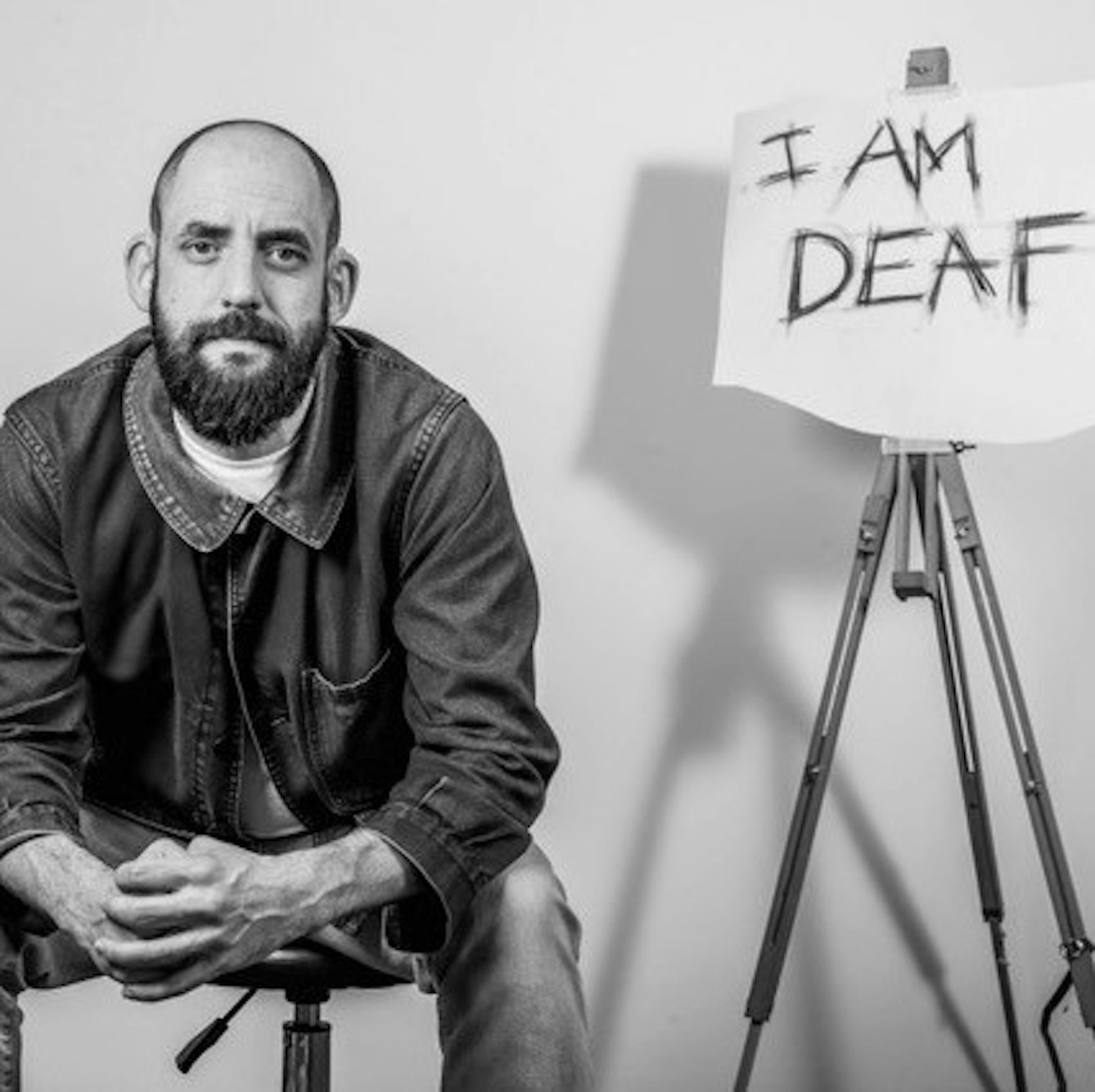 Black and white photograph of artist Jonny Cotsen. Sign in background reads "I am deaf"