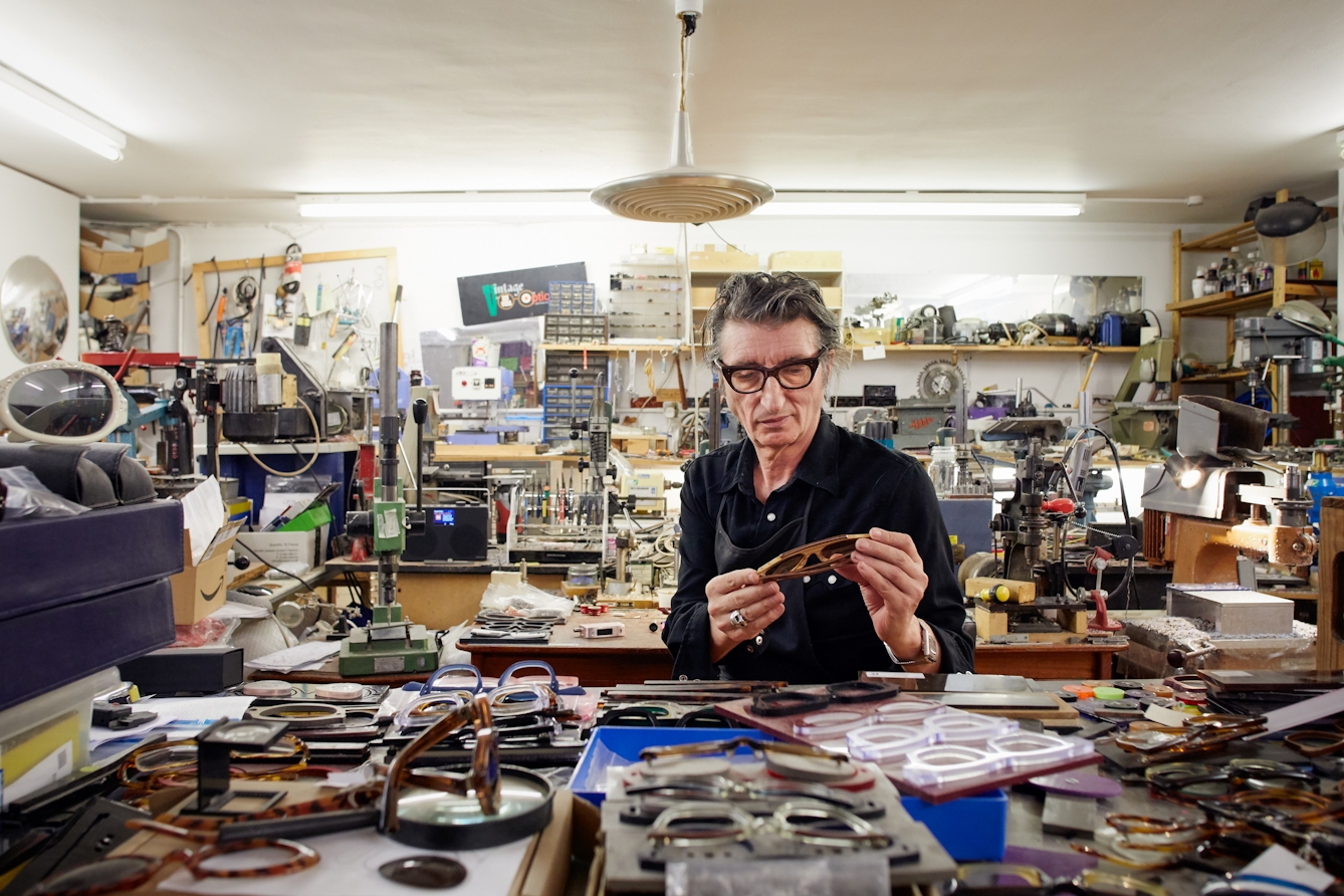 Photograph of a man in a black shirt wearing a pair of black framed spectacles, sitting in the middle of a cluttered workshop, surrounded by parts of spectacles, machinery, boxes and shelves. He is looking at a pair of spectacle he's holding in his hands.