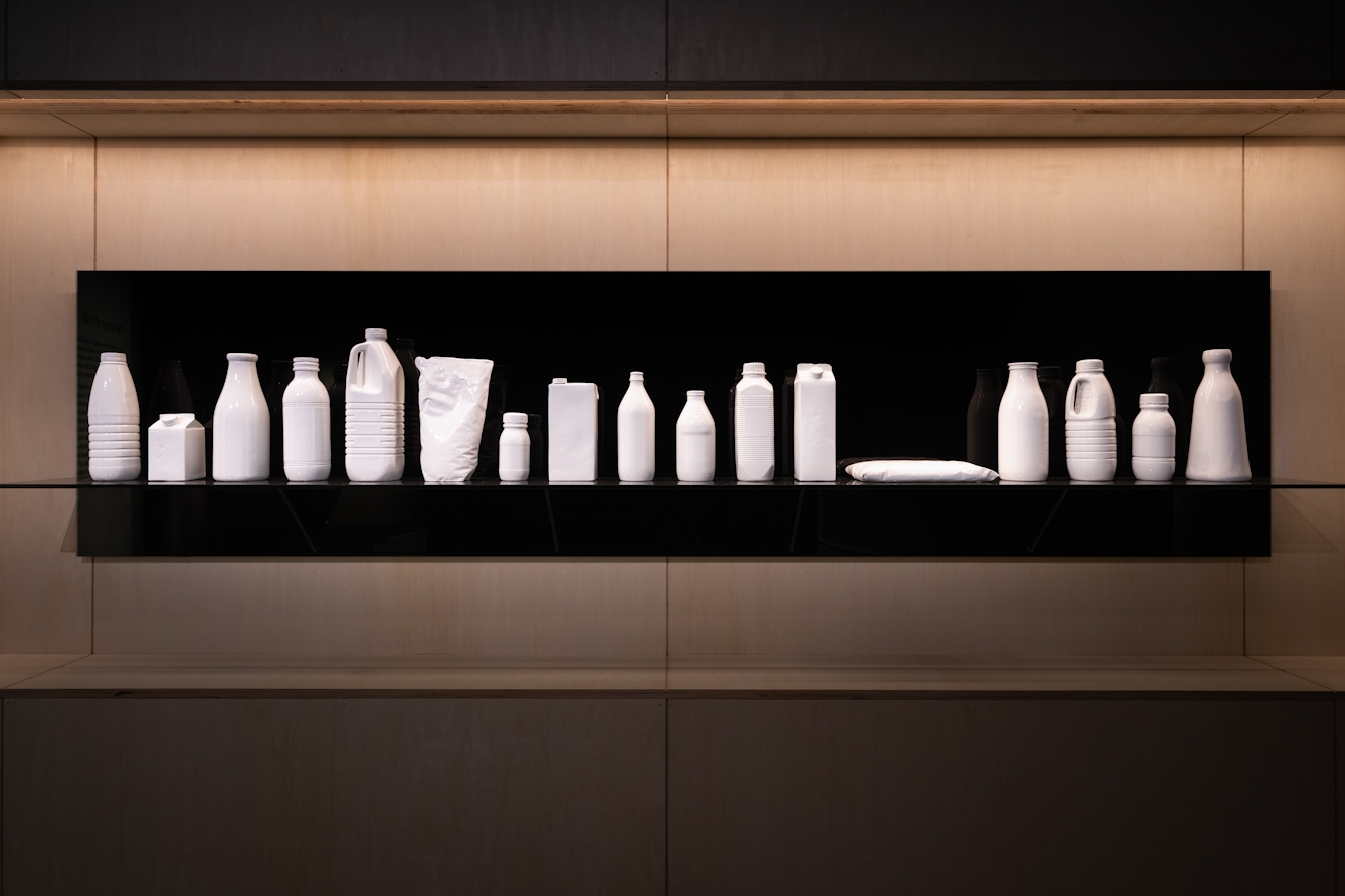 Photograph of a black, glossy perspex shelf with accompanying wall backing, hung on an exhibition gallery wall. Carefully placed on the shelf are 18 different white casts of milk bottles and packets from around the world.