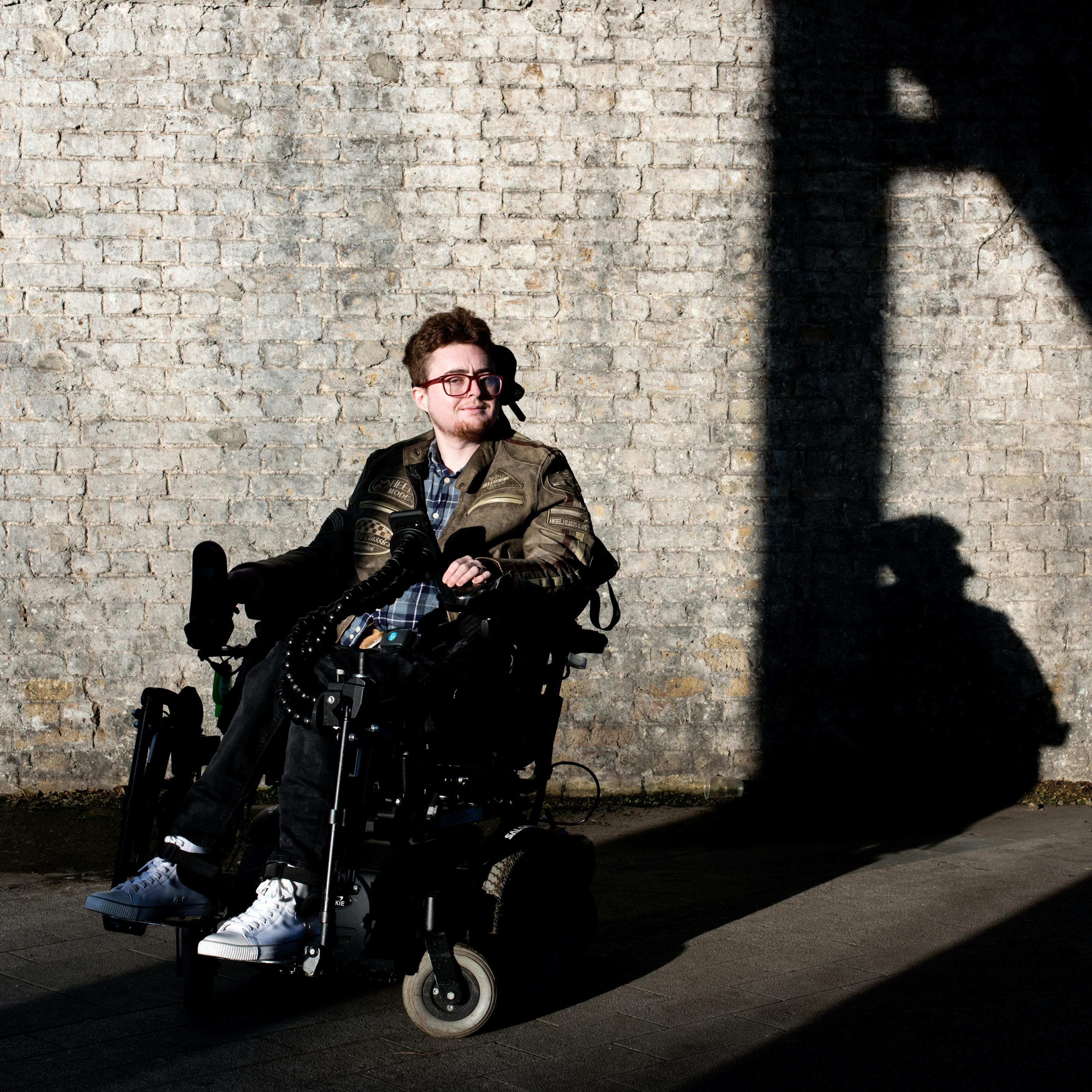 Photograph of a young person in a motorised wheelchair outside, in front of a white washed brick wall. They are lit by a low sun which is casting long shadows against the wall.