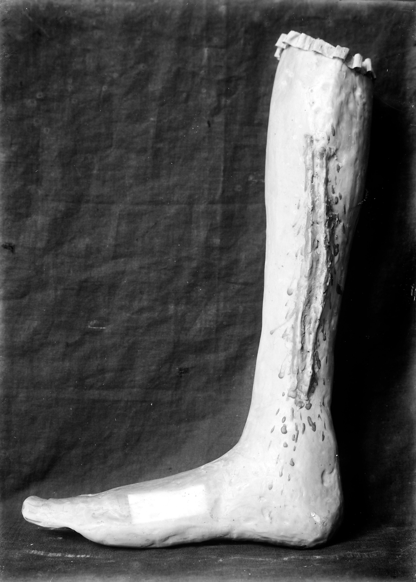 Black and white photograph of a wax votive offering. It is shaped like a human leg, starting from below the knee and finishing at the foot.  On the leg there is a visible scar and droplets of blood. There is also a lighter patch of skin on the top of the foot. 