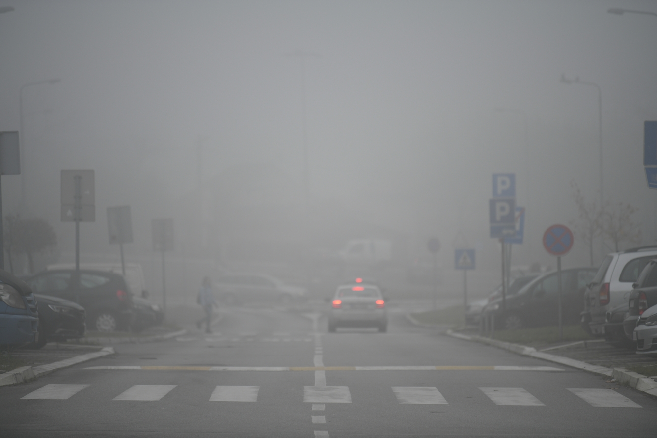 Colour photograph of a road and pedestrian crossing. There are parking signs on the left and right and parked cars to either side. In the left lane of the road is a white car that is driving, with its red tail lights on. It is very misty, and in the distance in the mist are several other cars and signs. 