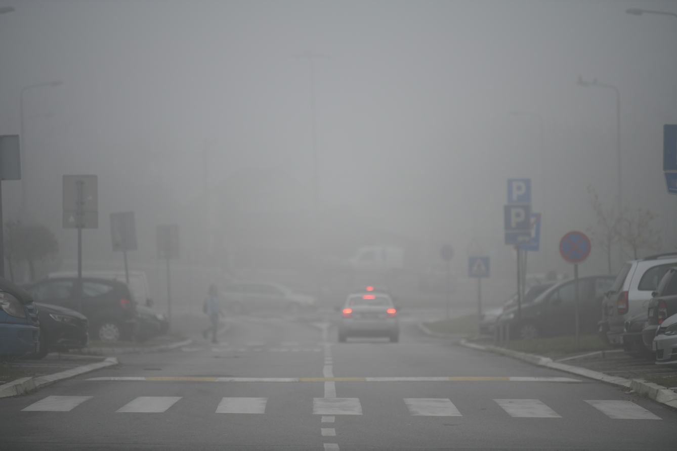 Colour photograph of a road and pedestrian crossing. There are parking signs on the left and right and parked cars to either side. In the left lane of the road is a white car that is driving, with its red tail lights on. It is very misty, and in the distance in the mist are several other cars and signs.