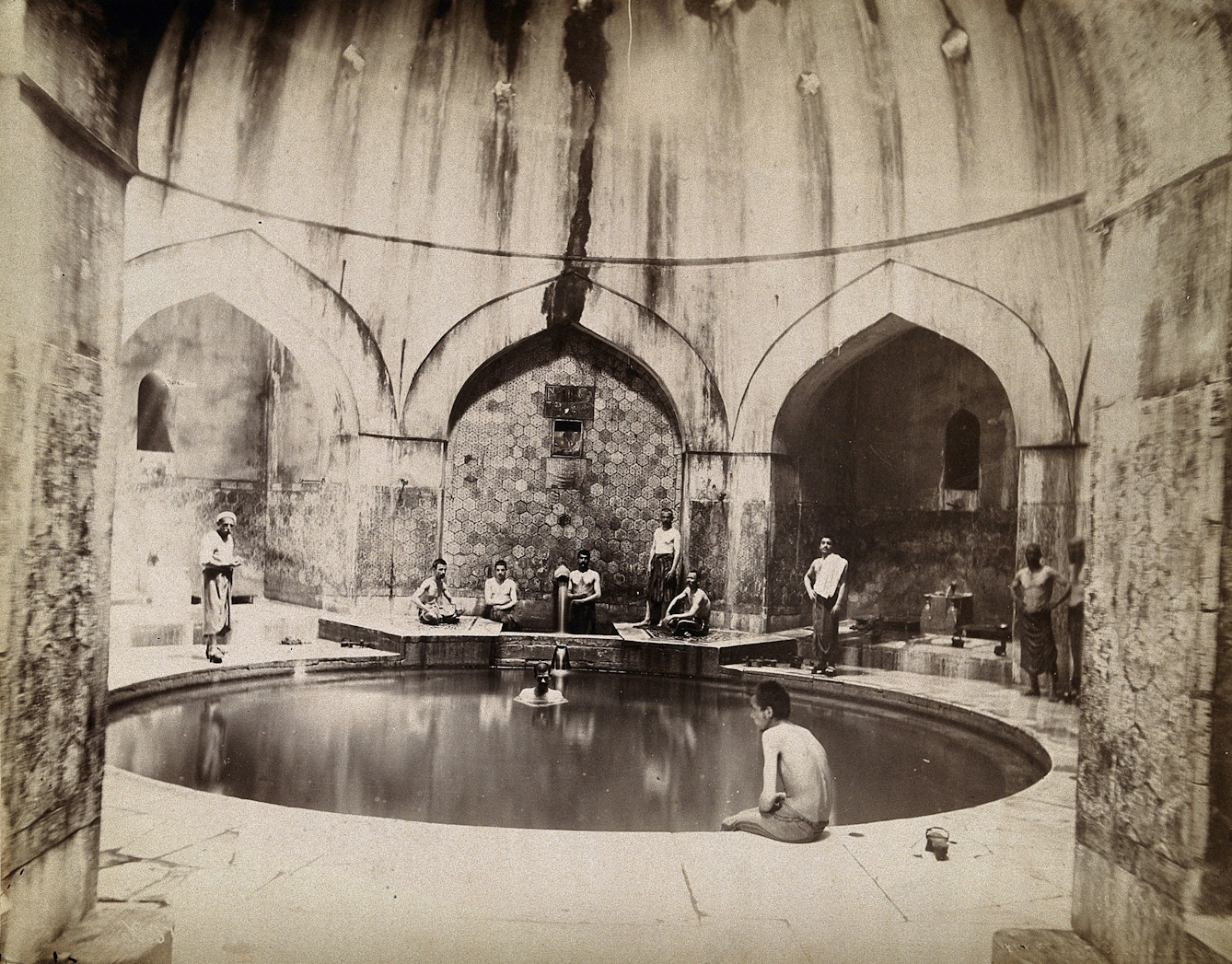 Black and white photograph showing men sitting around the sides of a circular pool in a domed building.