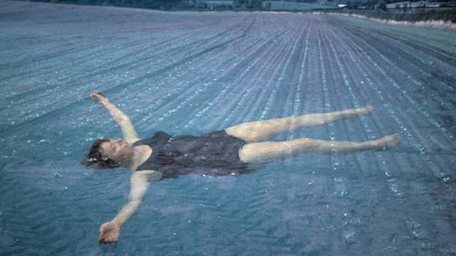 Photograph of Jo Spence wearing a black one-piece swimming costume floating face up in blue rippled water