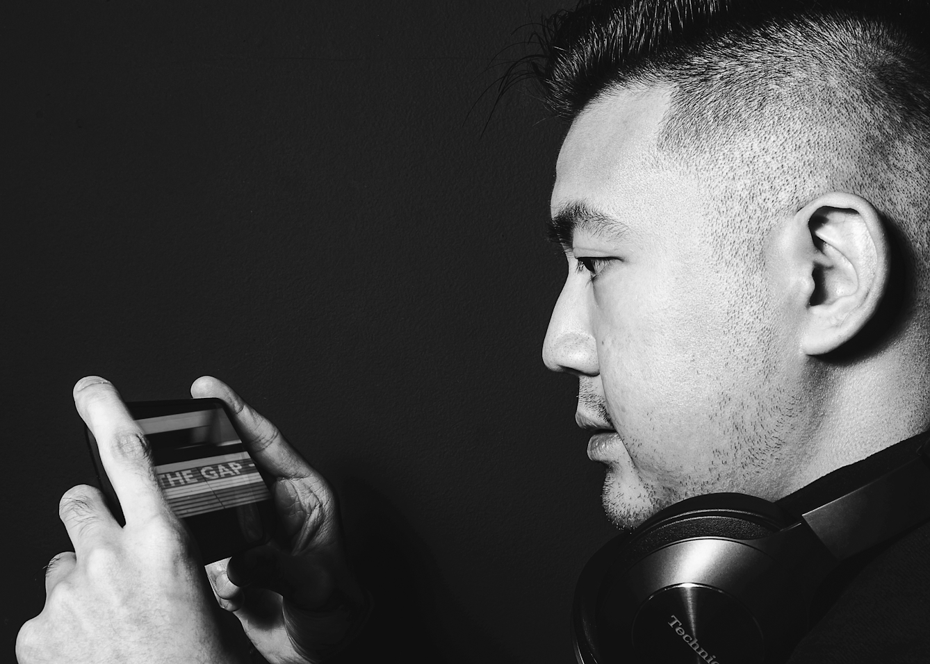 Black and white photographic portrait of a close-up of a man's face in profile. He is holding up a smartphone in his hands. His eyes are directed towards the phone's screen. He has a set of over ear headphones resting on his shoulders around his neck. The man's face and hands are spotlit in a small circle of light. He is standing against a black background which means he is surrounded by darkness. On the screen of the phone is an image of the edge of a train platform with the large words, 'The Gap'.