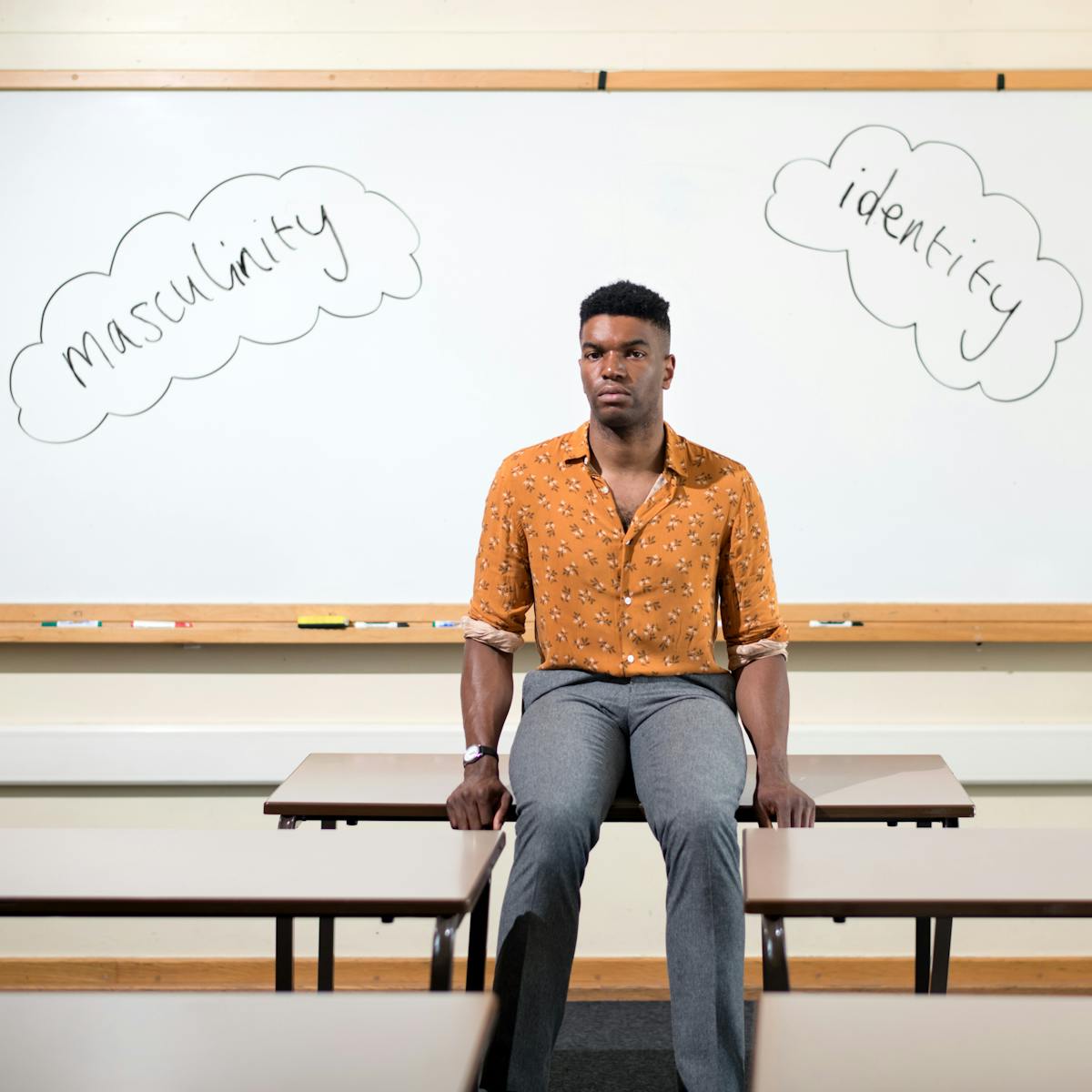 Photograph of a man in an orange shirt and grey trousers sat on a desk in a classroom. behind him on the white board is written the words, 'masculinity' and 'identity'.