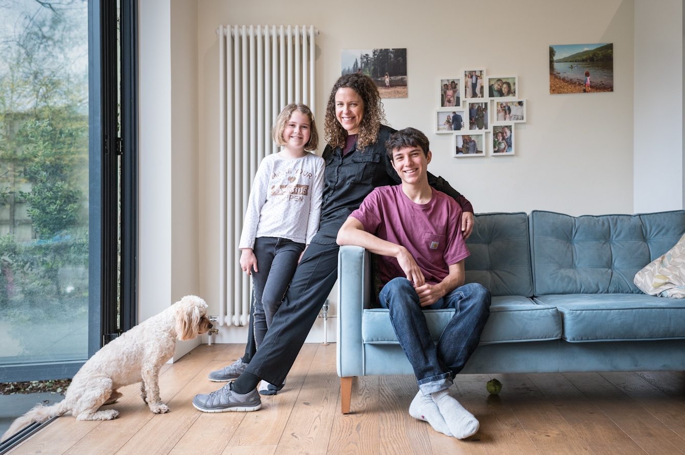 Photograph of a mother and her two children in their living room. They are sat on the sofa and the mother is in the middle, her arm around each one. In the background are family photos in frames hung on the wall. At the family's feet is the family dog.