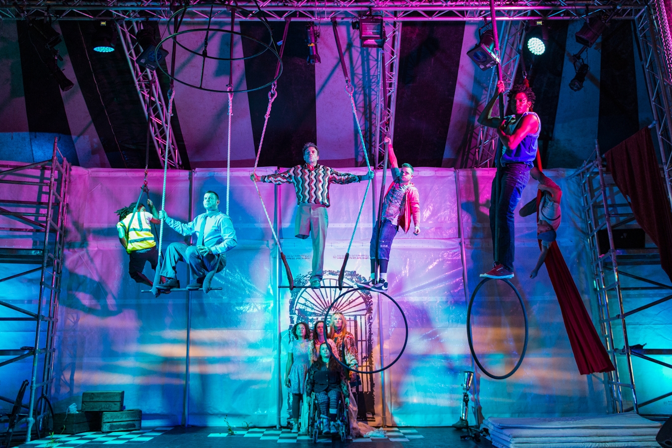 A wide angle photograph of a theatrical production lit with blue and purple lighting. A group of five performers are standing on bars and hoops suspended from the ceiling. The performer standing on the central ‘trapeze’ has his arms outstretched, holding the ropes. His right trouser leg is hemmed just above the knee revealing that he does not have a right leg. To the centre-bottom of the frame a further group of three performers are standing over the shoulders of a performer seated in a wheelchair.