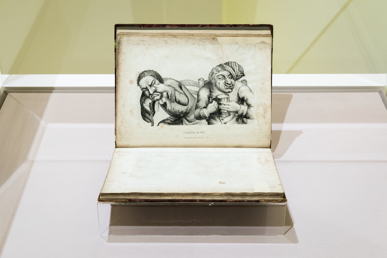 Photograph of a glass exhibition display case showing an open printed book, held in a book cradle. The book is in landscape orientation and on the upper page is an etching of two characters, one crying and rubbing their eyes and the other holding a flagon of beer and smiling.