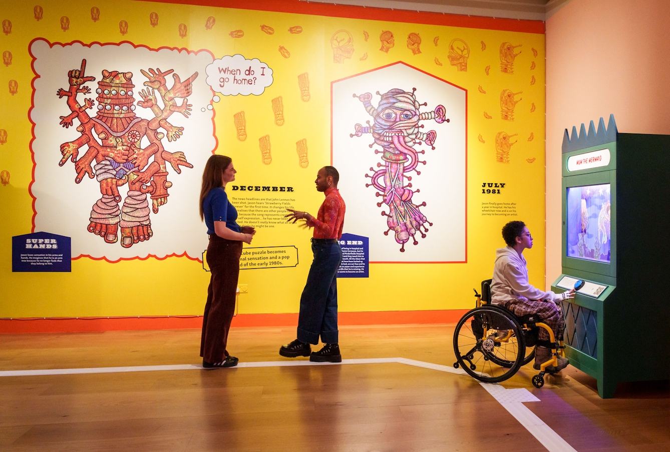 Photograph of two people standing in front of a wall. The wall has brightly coloured wallpaper with drawings and words on it.