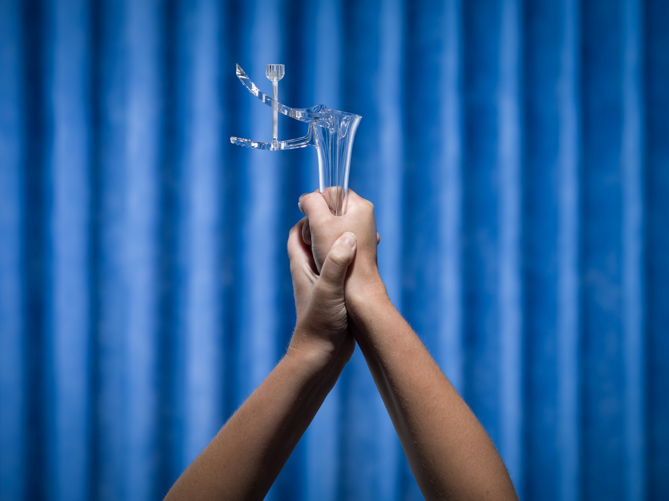 Photograph of a pair of hands firmly holding shut a vaginal speculum. Photographed against a background of blue antibacterial clinical curtains.
