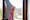 Photograph of a woman wearing a pink head scarf. We see a fragment of her face reflected in a shard of mirror. behind the mirror is a window and the view of the out of focus outside.