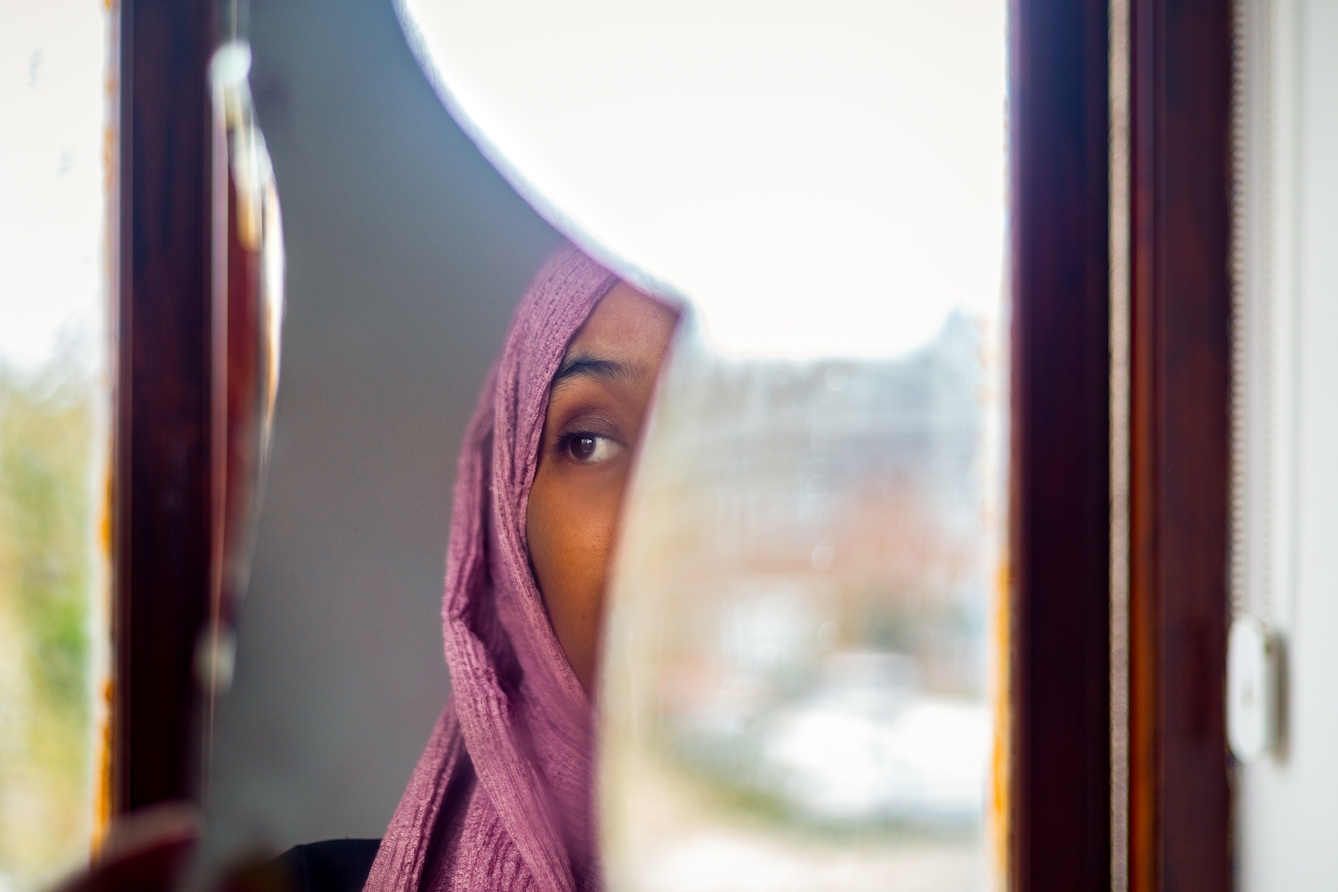 Photograph of a woman wearing a pink head scarf. We see a fragment of her face reflected in a shard of mirror. behind the mirror is a window and the view of the out of focus outside.