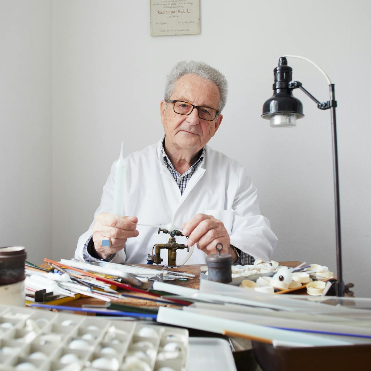 Photograph of an older man in a white lab coat siting at a work table in a domestic house. He is holding white glass tubing. In front of him on the table is a small gas burning torch, glass rods, a box of glass eyes and a small lamp on a stand.