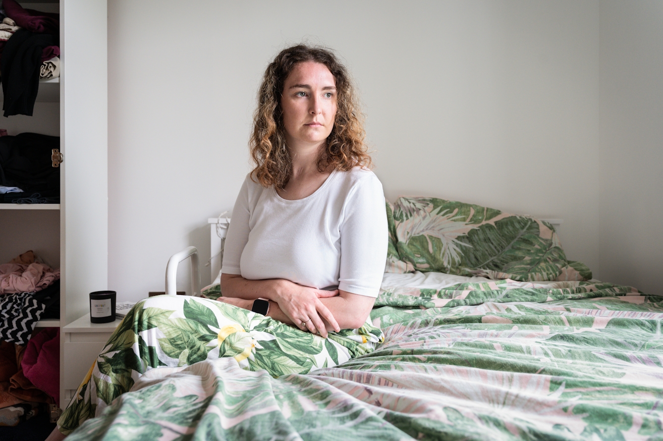 Photograph of a woman with shoulder length frizzy brown hair, wearing a white elbow length top and leaf patterned trousers, sitting on the side of a double bed. Her arms are loosely folded across her lap. She is looking off in the distance to camera right towards the light from a window. She has a calm neutral expression. The duvet cover and pillow covers are also have a green leaf pattern, echoing her trousers. Behind her the wall is white and visible behind her on the left hand side is a small white bedside table and an open fronted wardrobe with clothes folded on the shelves.