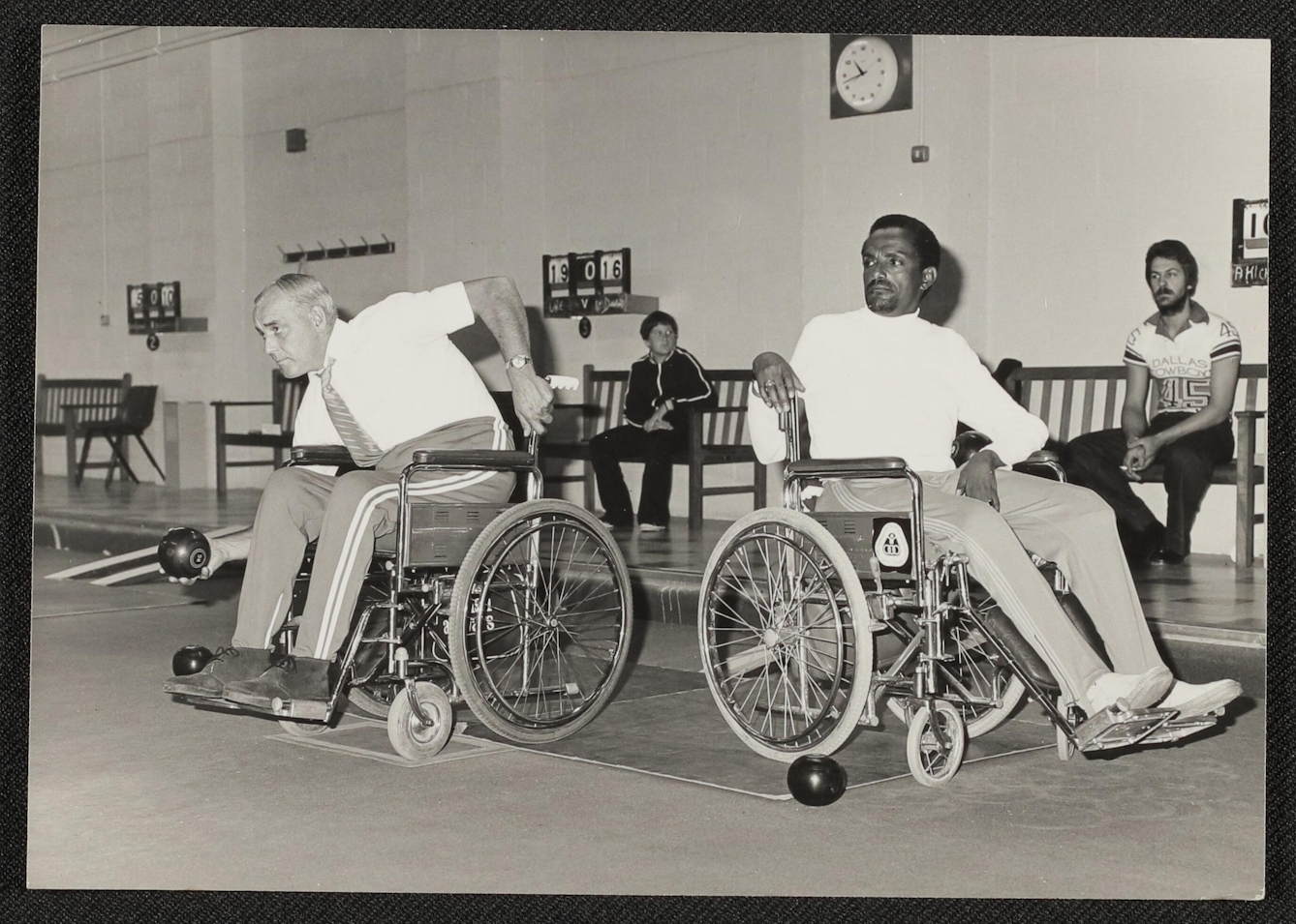 Black and white photograph showing two men playing wheelchair skittles. On the left, a white man leans down, gripping his wheelchair for balance using his left arm and reaching forward with a black skittle in his right hand, about to throw it. To his right sits a black man with his wheelchair pointing away from the throwing man, who is leaning around in his chair to watch. Behind are benches for spectators, one of whom is a bearded man in a Dallas Cowboys shirt holding a cigarette, and the other of which is a teenage boy in a dark tracksuit with white stripes running down the sleeves and front. A scoreboard in the background appears to indicate the score is 19:16. 