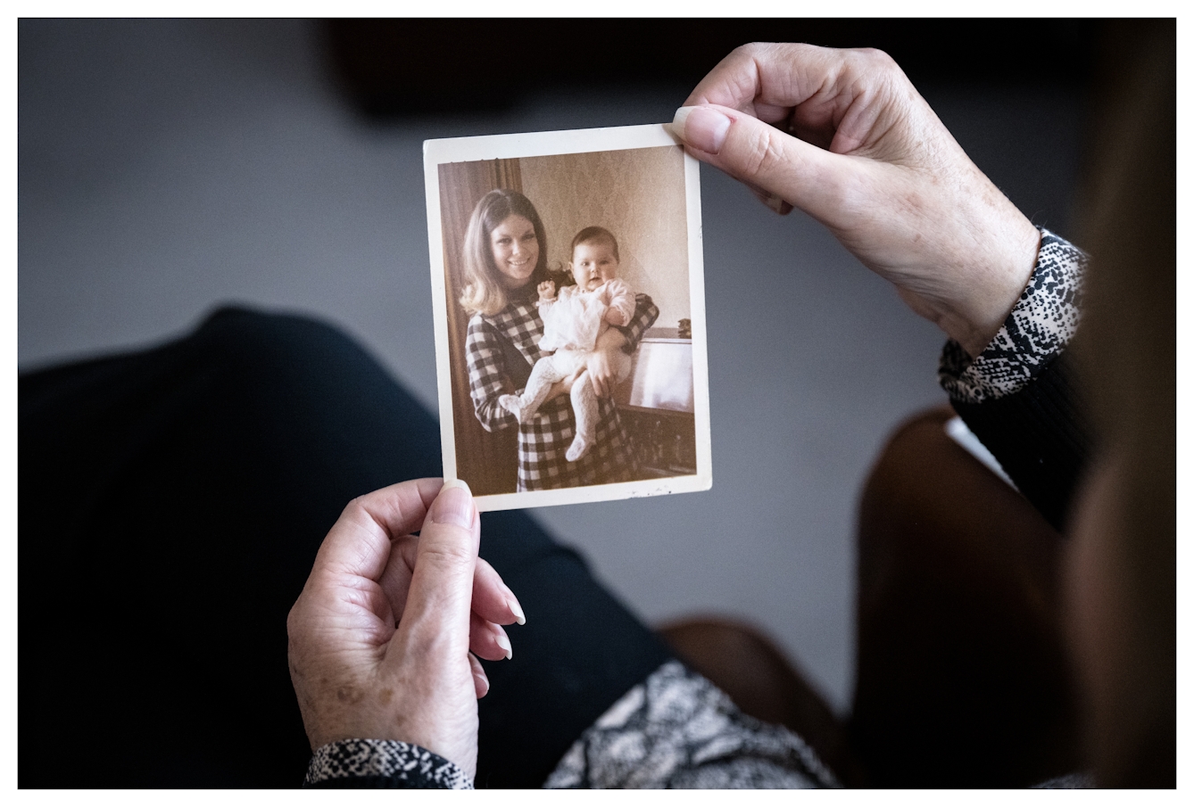 Photograph of Marie Lyon's hands holding an old photograph of her when she was younger holding her baby daughter in her arms. The young Marie is smiling to camera. Her daughter left arm isn't fully formed and the tiny palm and fingers that had grown where it stopped, at her elbow are visible.