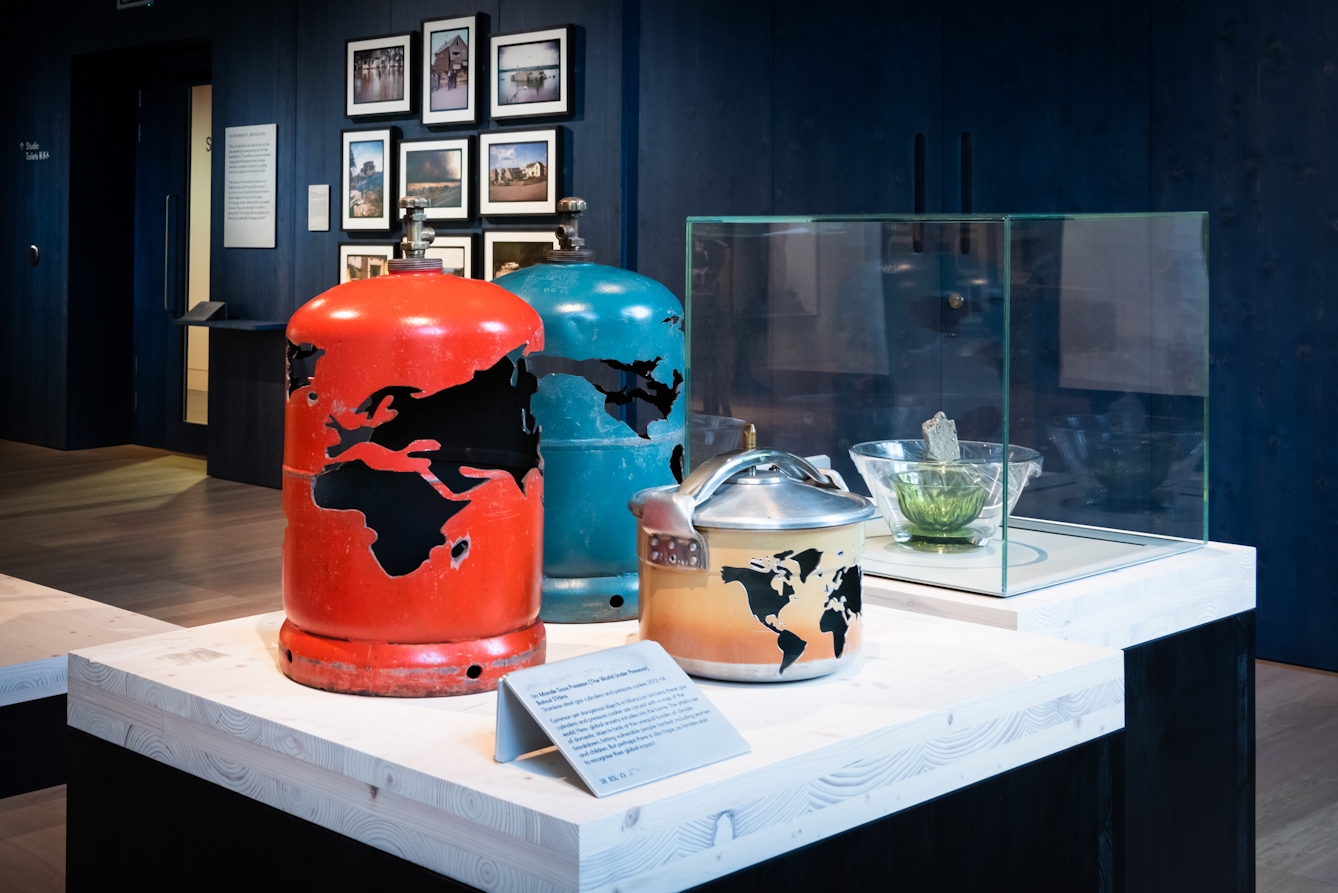 Photograph of an artwork on a display plinth in a gallery space. The artwork consists of 2 gas canisters and a pressure cooker, each one has a map of the earth's landmasses carved out of its casing.