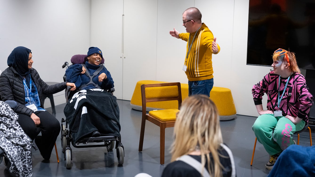 A group sitting in a circle at an event in a studio space with white walls. A student in a wheelchair is smiling at a man who is standing speaking to the group, he is wearing a yellow jumper and glasses, he is holding his arms out wide and engaging with the student. 