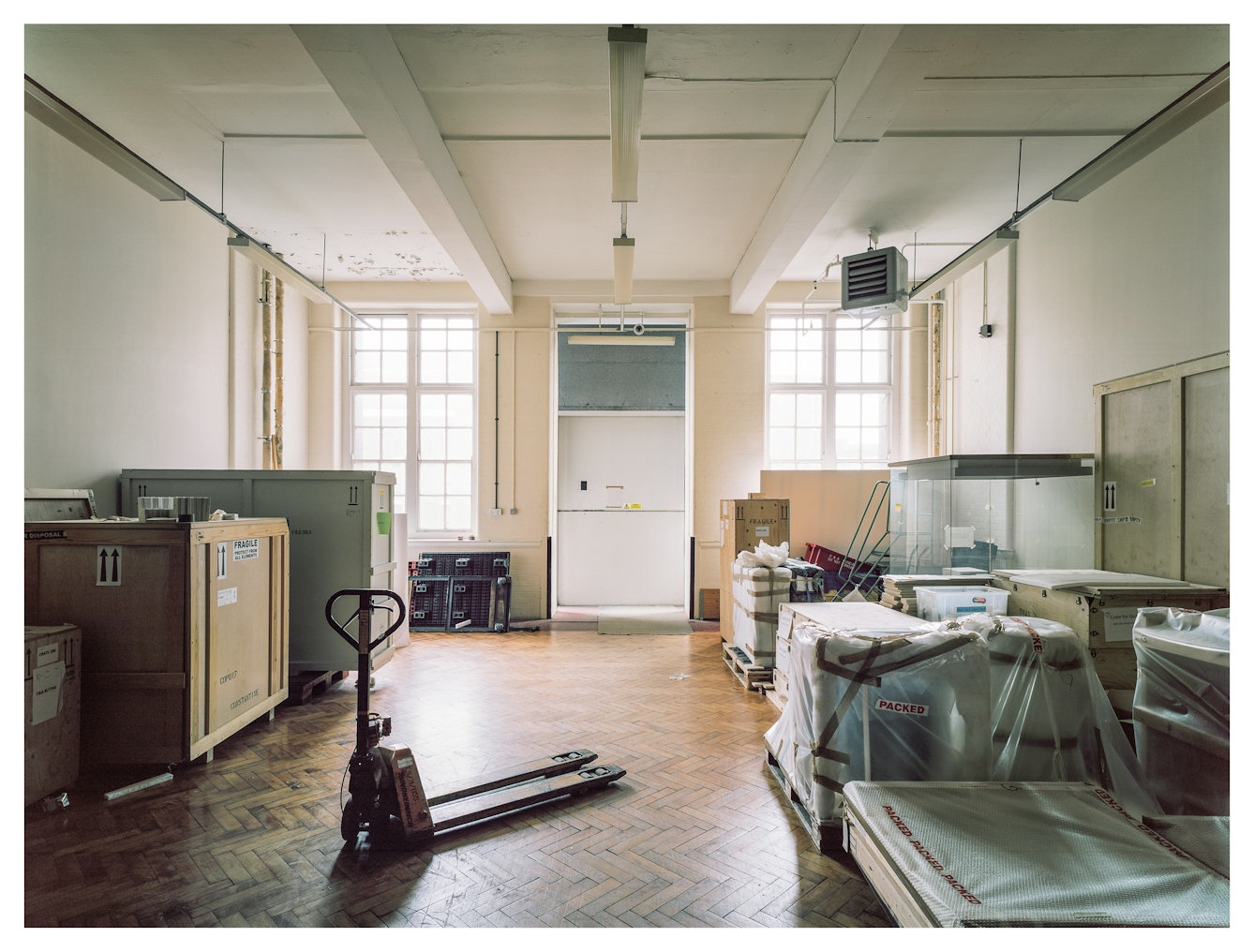 Photograph of a large room, with two large windows though which daylight is flooding. The floor is wooden parquet. Neatly arranged in the room are large wooded crates, empty glass display cases and plastic wrapped objects. At an angle in the foreground is a pallet lift.