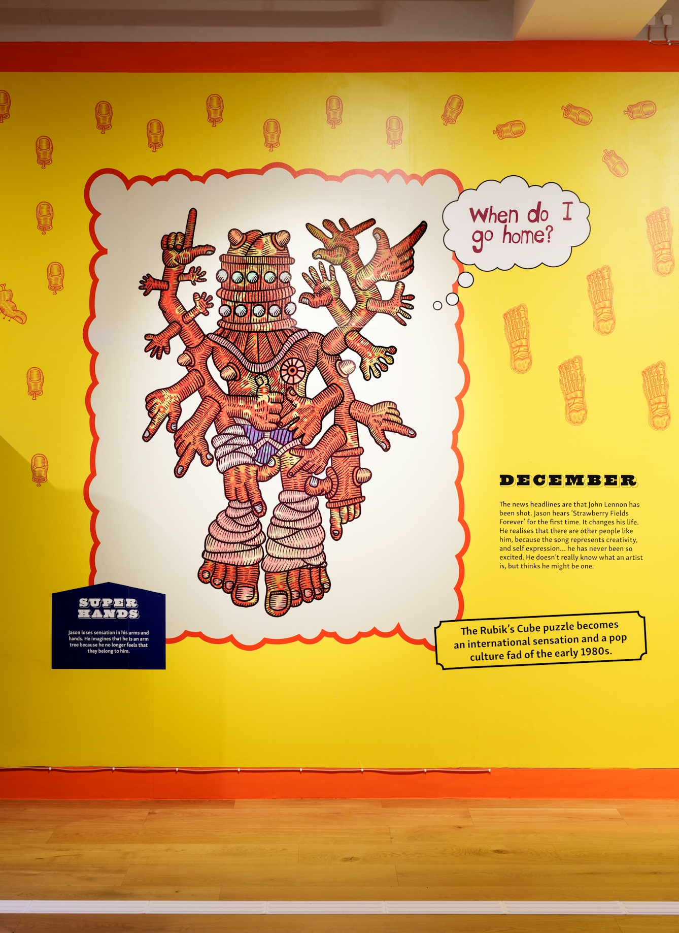 Photograph of a colourful yellow wallpaper mural covering an entire gallery wall. The mural contains text and drawings. This section show a multi-handed character pointing in all directions. The text beside it is titled 'December'.
