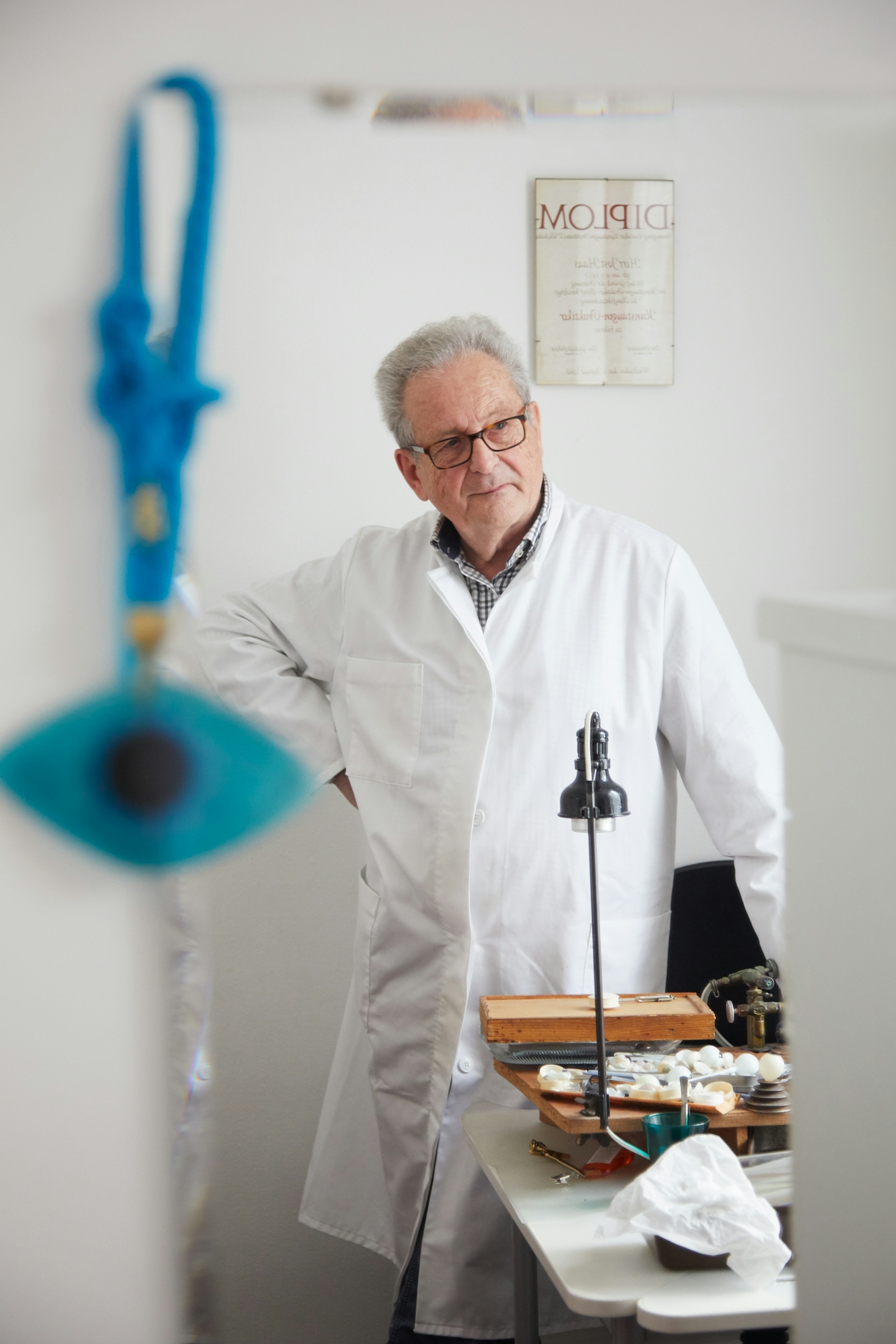 Photograph of an older man wearing a white lab coat in the reflection of a mirror He is standing at a workbench with covered in tools and glass eyes, with his hand on his hip, looking off into the distance. Hung on the corner of the mirror is a blue eye on a blue cord.