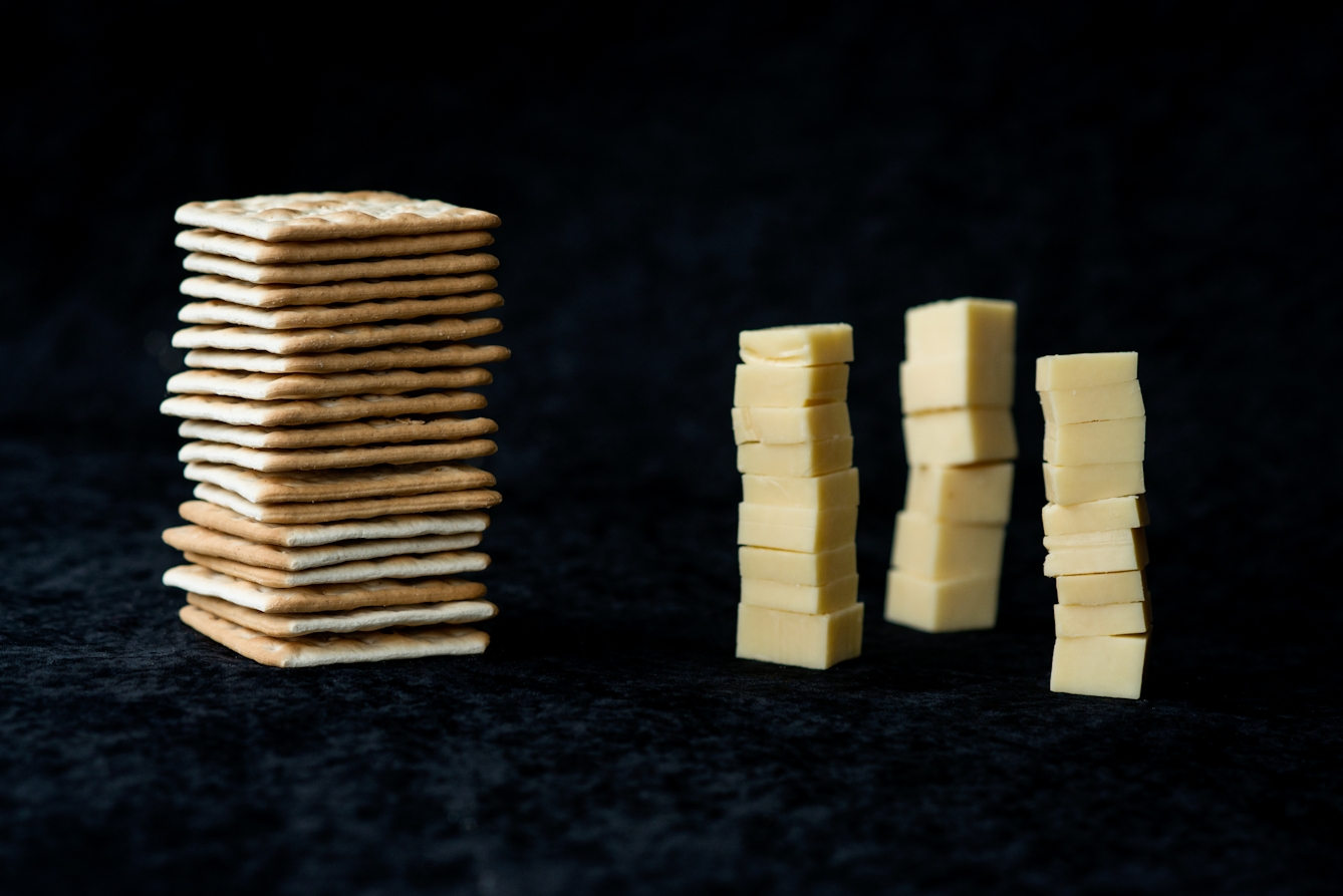 Photograph of a pile of cheese crackers on the left and three towers of cubed cheese on the right. The background is a black velvet material.