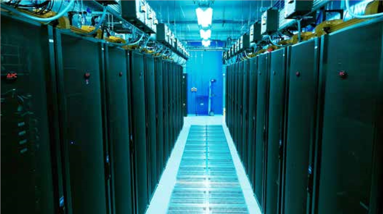 A long room filled with two rows of computer processing equipment in the form of rectangular stacks to overhead cables. A corridor extends between the two stacks of computers.