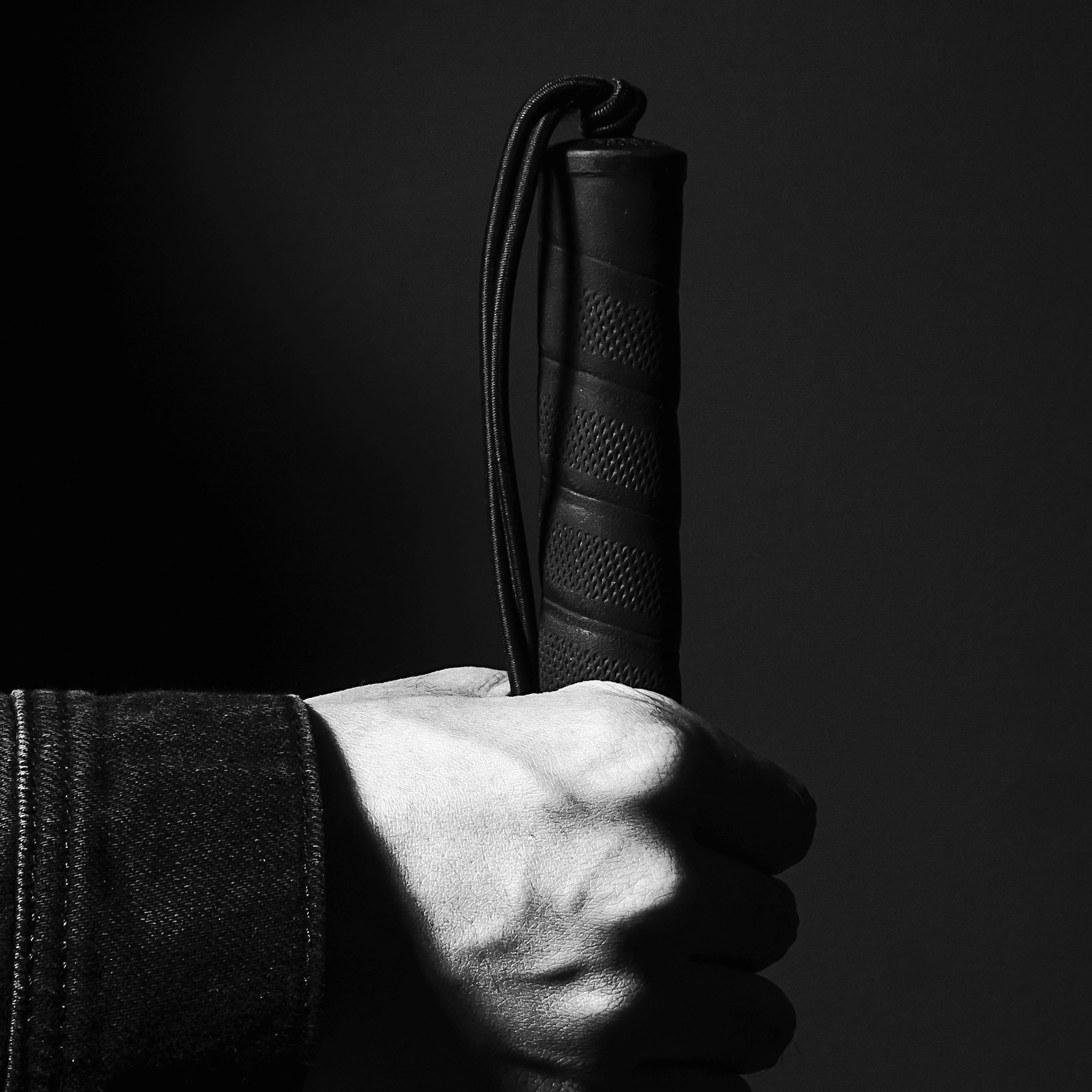 Black and white photograph of a hand holing the rubber handle of a white cane such that the cane is vertical. The image is close-up on the hand so that all that can be seen is the cuff of the person's denim jacket. The background is a dark grey making the hand and handle a stark feature within the image.