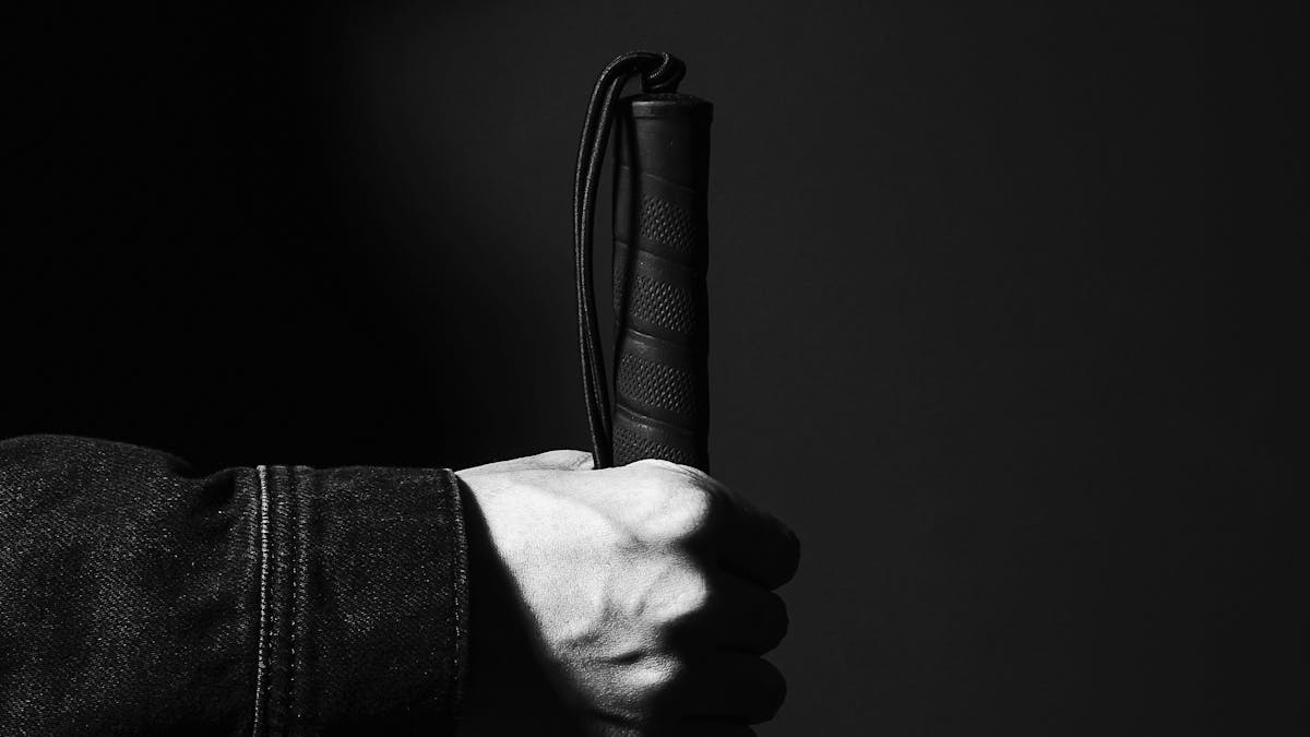 Black and white photograph of a hand holing the rubber handle of a white cane such that the cane is vertical. The image is close-up on the hand so that all that can be seen is the cuff of the person's denim jacket. The background is a dark grey making the hand and handle a stark feature within the image.