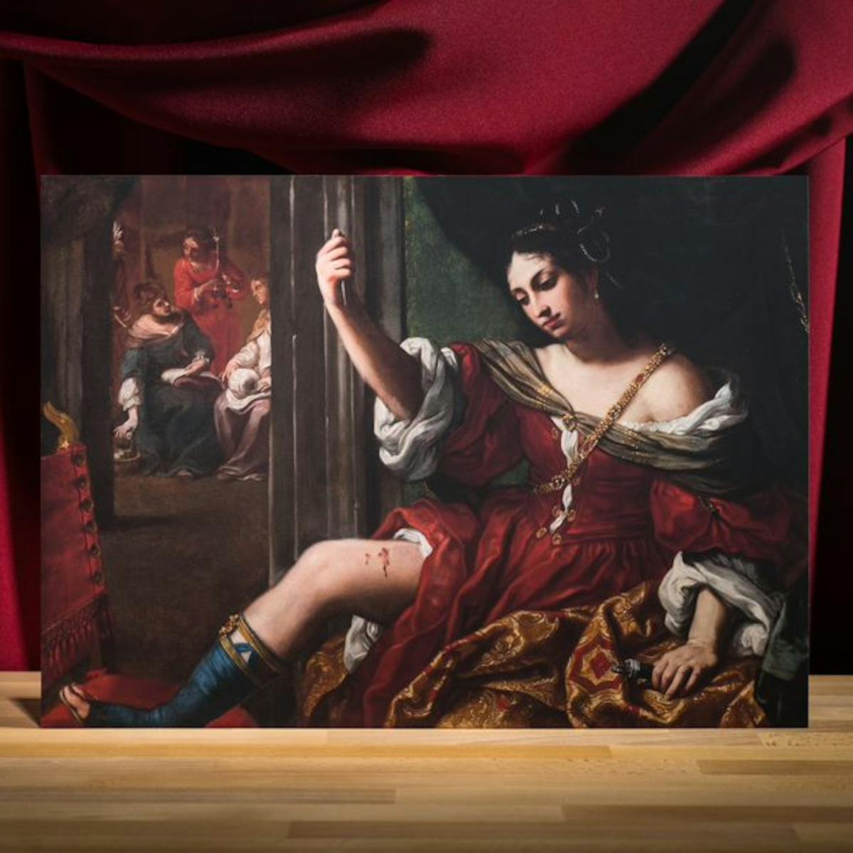 Photograph of an oil on canvas painting of a woman raising her hand and holding up a knife towards her exposed thigh. Her thigh is wounded and blood is coming out of the wounded sight. She is wearing a grand red dress and a grand looking necklace. 