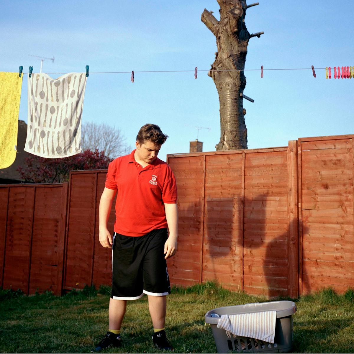 Photograph of a teenage boy wearing a red t-shirt and black shorts, standing in a garden. Above him is a washing line with pegs and towels.