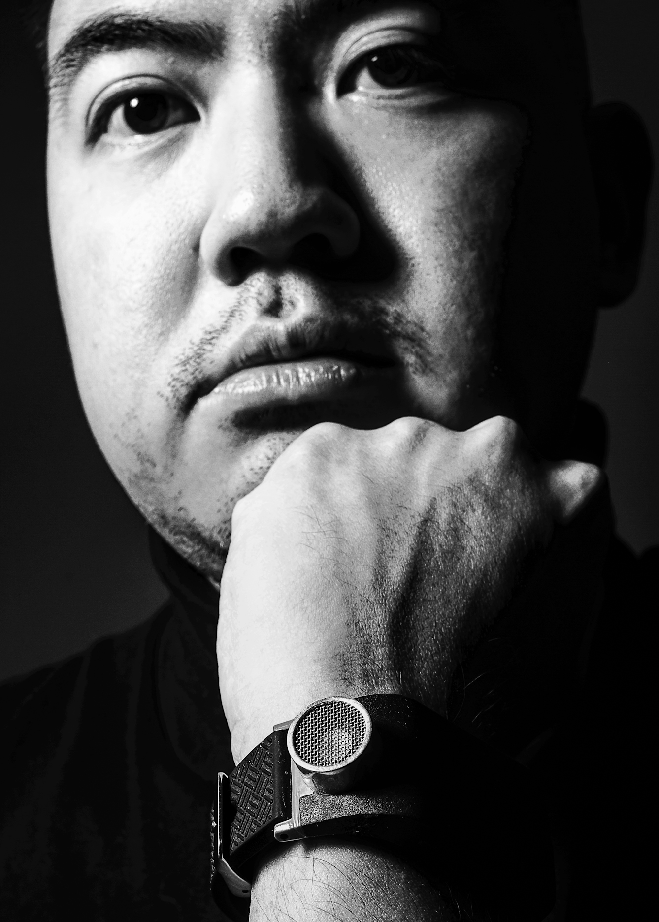 Black and white photographic portrait of a close-up of a man's face looking straight to camra. His chin is resting on his left hand. Around his wrist is a circular device with a mesh front attached to a wide leather strap, it is not a watch. The man's face and hand are spotlit in a small circle of light. He is pictured against a black background which means he is surrounded by darkness.