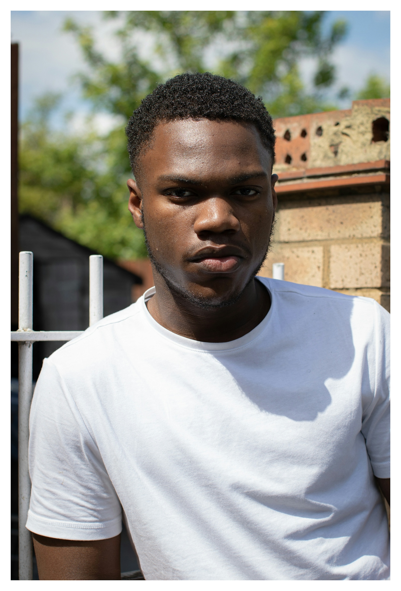 Photographic portrait of a young Black man wearing a white t-shirt. He is looking to camera leaning slightly on his right arm. He has a neutral but engaging expression. Behind him are out-of-focus elements of a garden scene.
